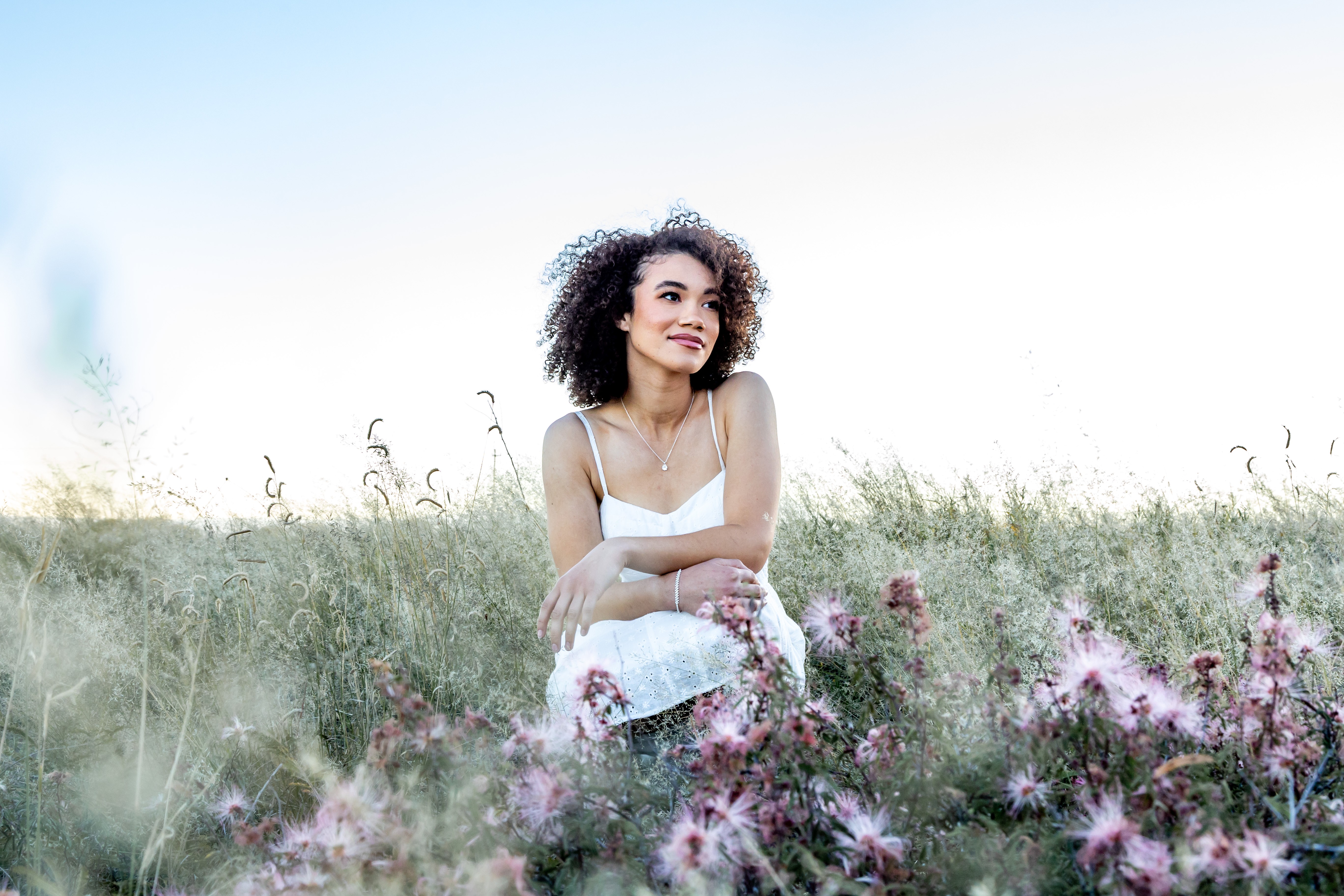 Young woman with curly hair sitting in a field of pink flowers, smiling and looking to the side.