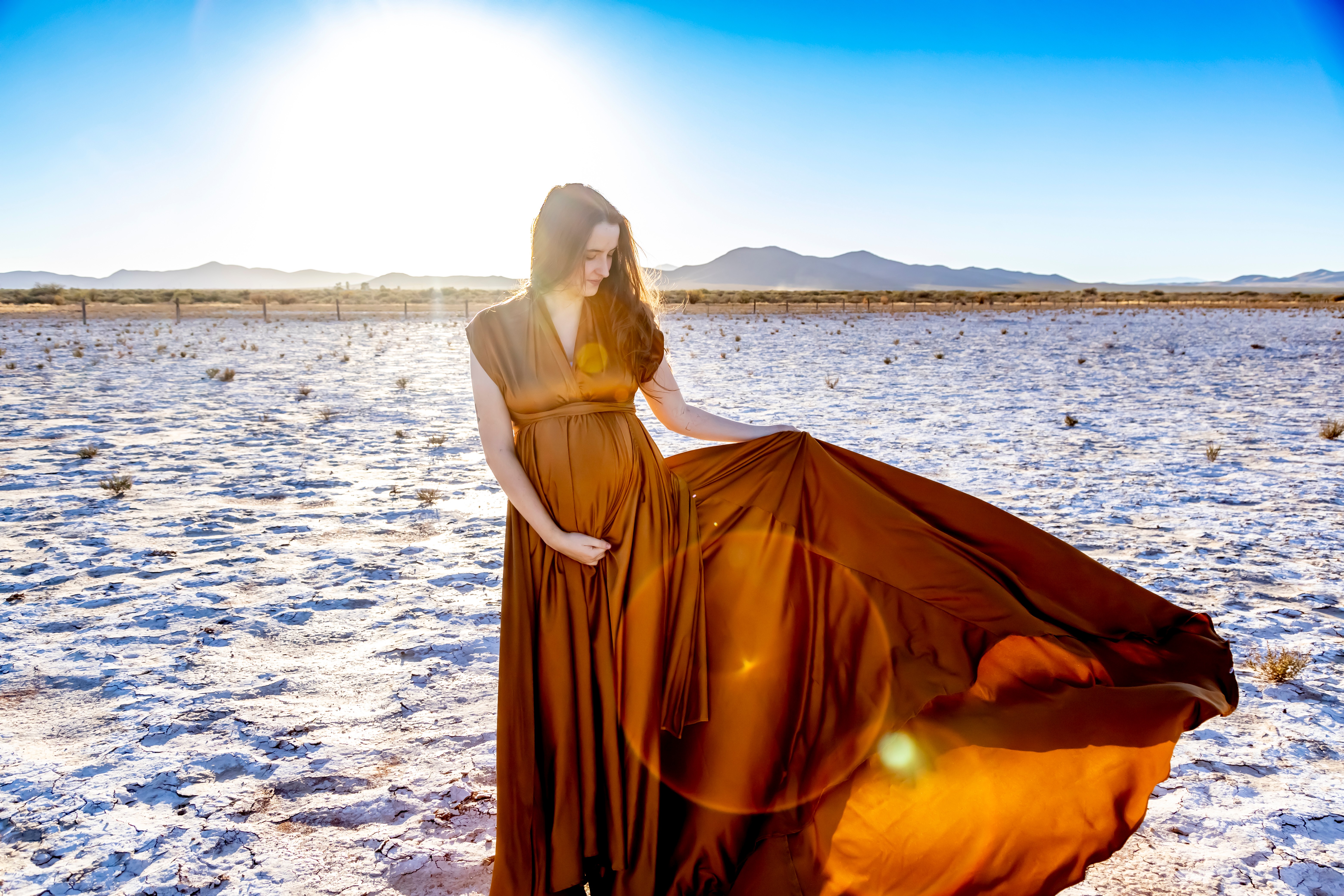 Woman in a long brown dress standing on a dry, flat landscape with mountains in the distance and bright sunlight.