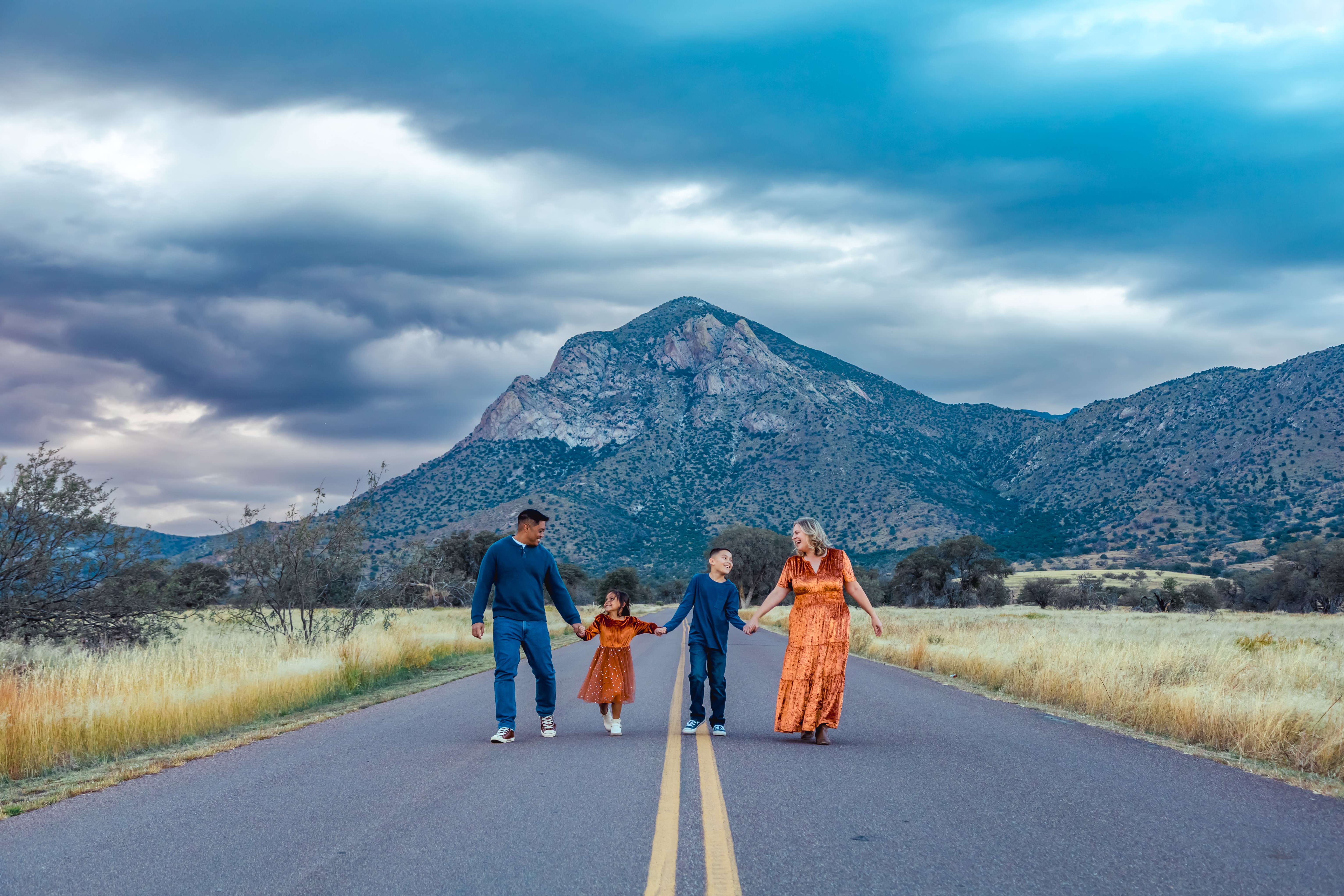 Family of four walking on a road with mountains and cloudy sky in the background.