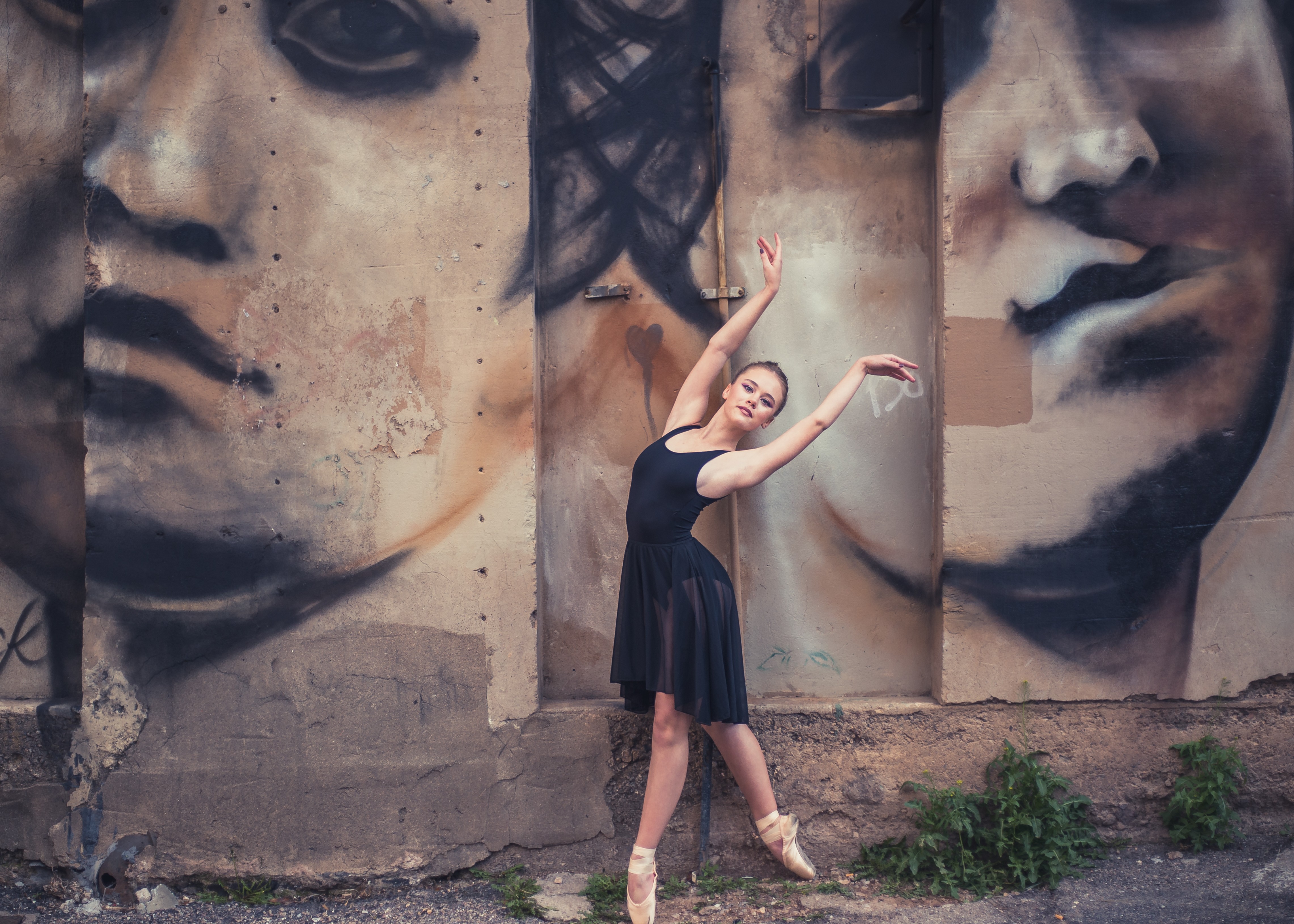 Ballet dancer in black dress poses in front of large mural of faces on wall, with plants at base.
