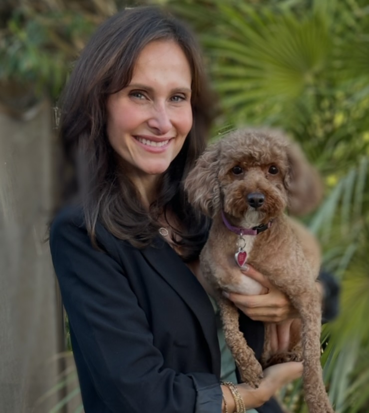 Woman holding a small curly-haired dog outdoors with green plants in background.