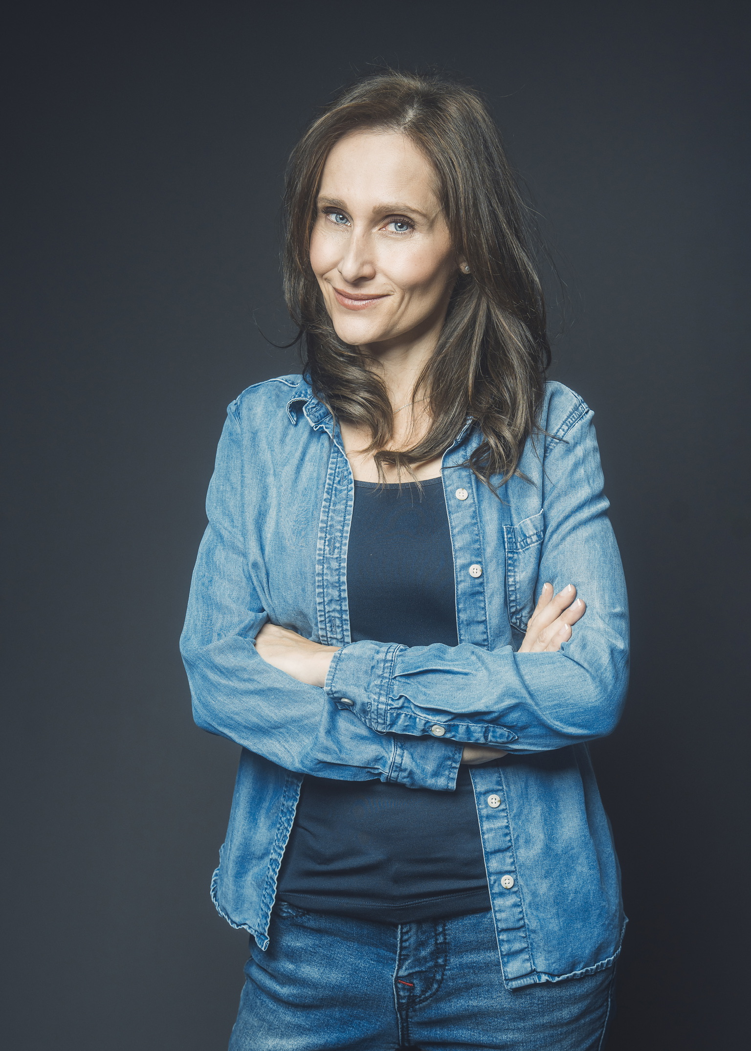 Woman with shoulder-length brown hair, smiling, wearing a denim jacket and dark shirt, arms crossed, dark background.