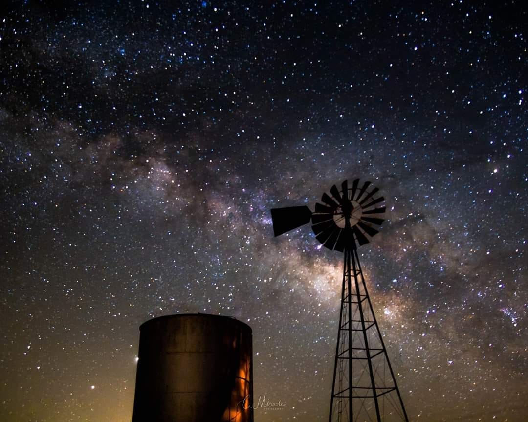 Night sky filled with stars and the Milky Way galaxy, with a windmill and a water tank silhouette in foreground.