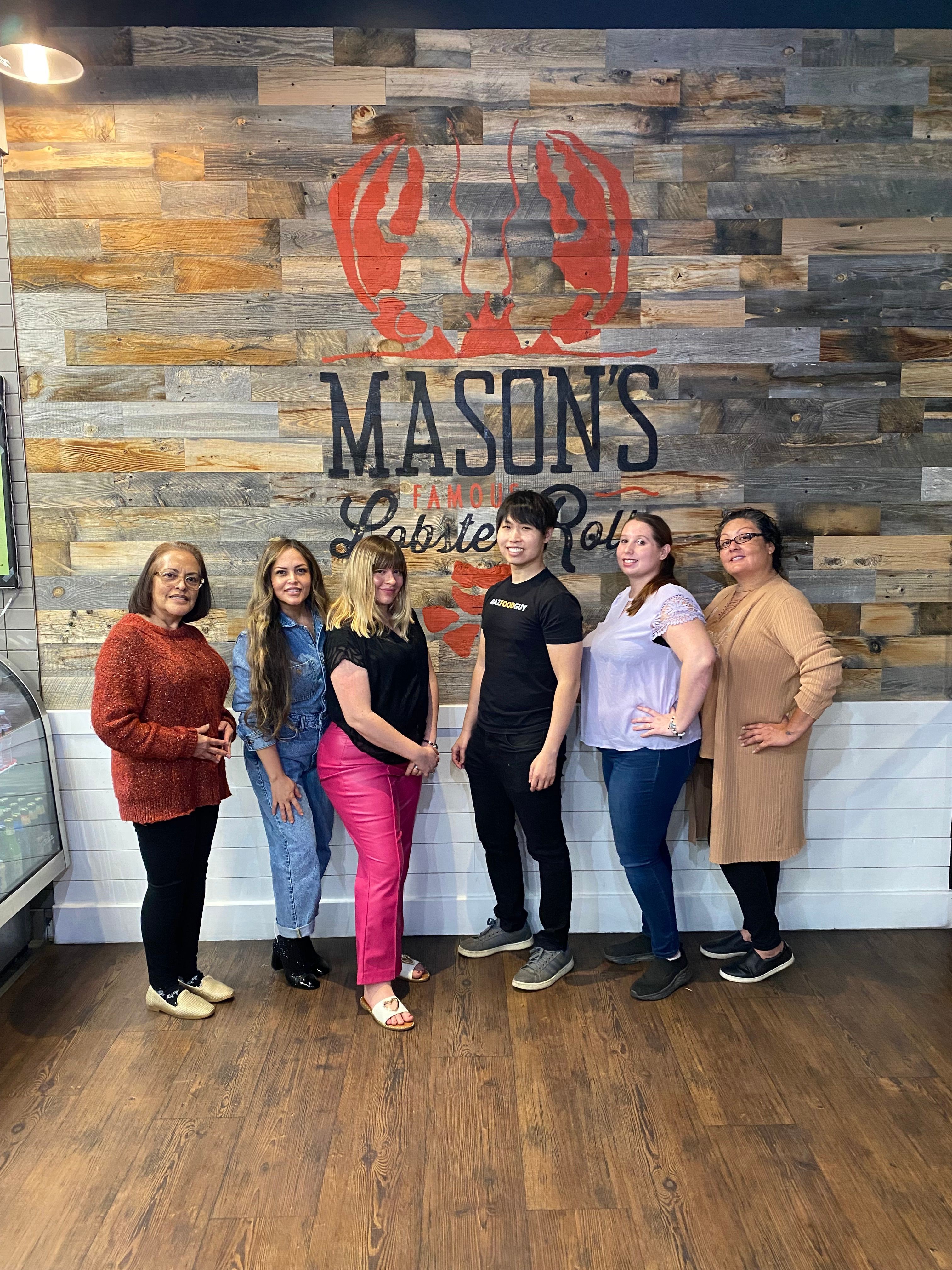 Group of six women standing indoors in front of a Mason's logo wall, smiling and posing for the photo.
