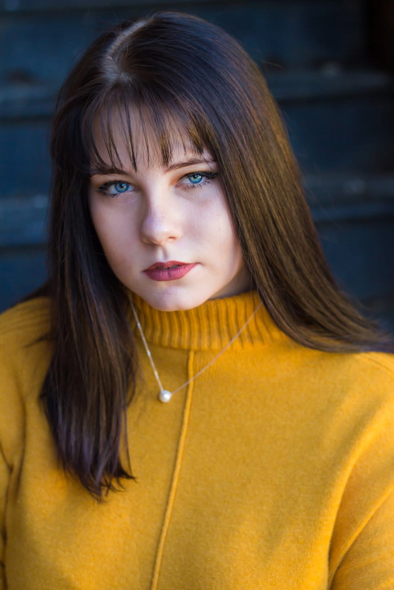 Young woman with long dark hair and blue eyes wearing a yellow sweater and a pearl necklace.