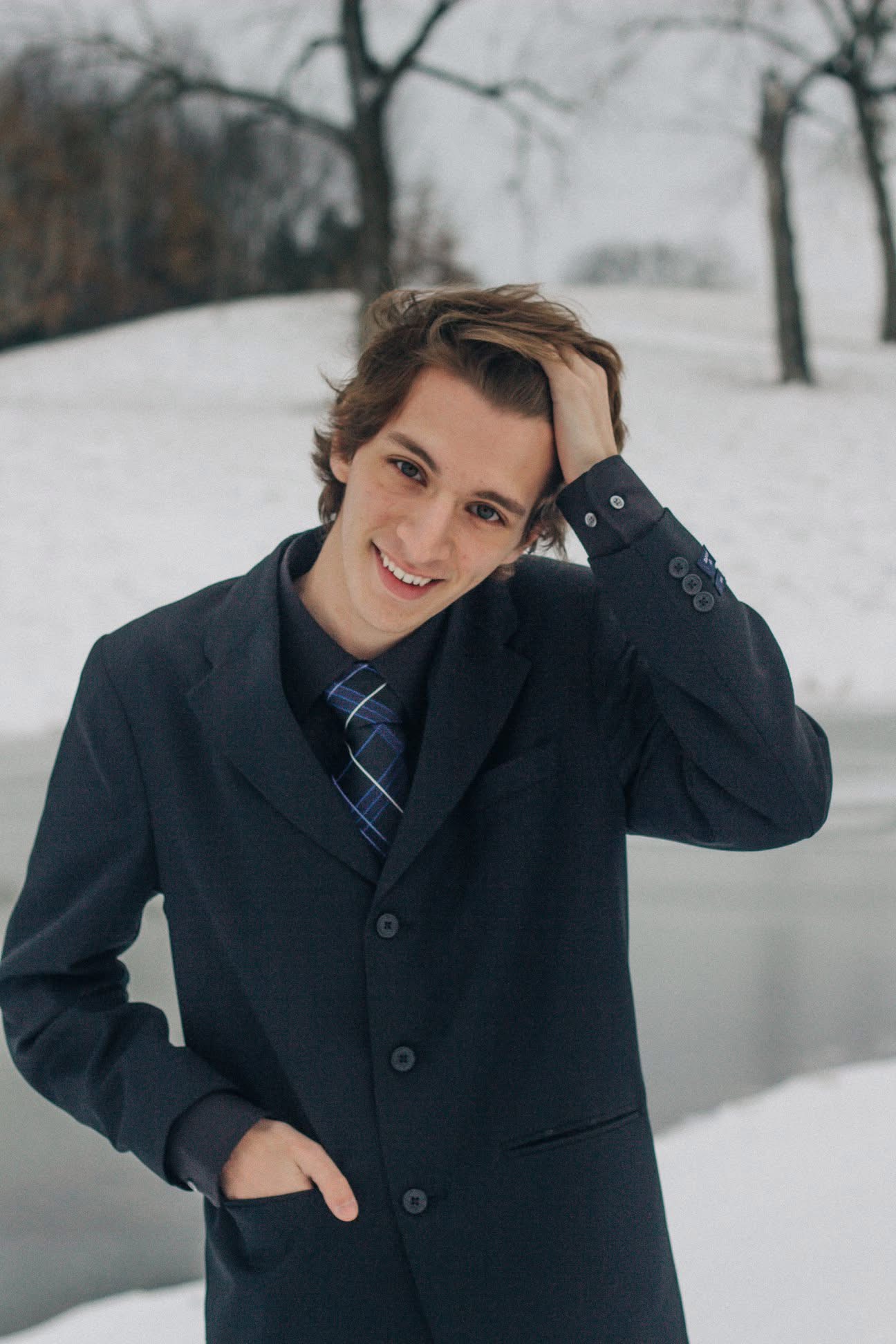 Young person with short hair in a dark suit and tie, smiling outdoors in a snowy landscape.