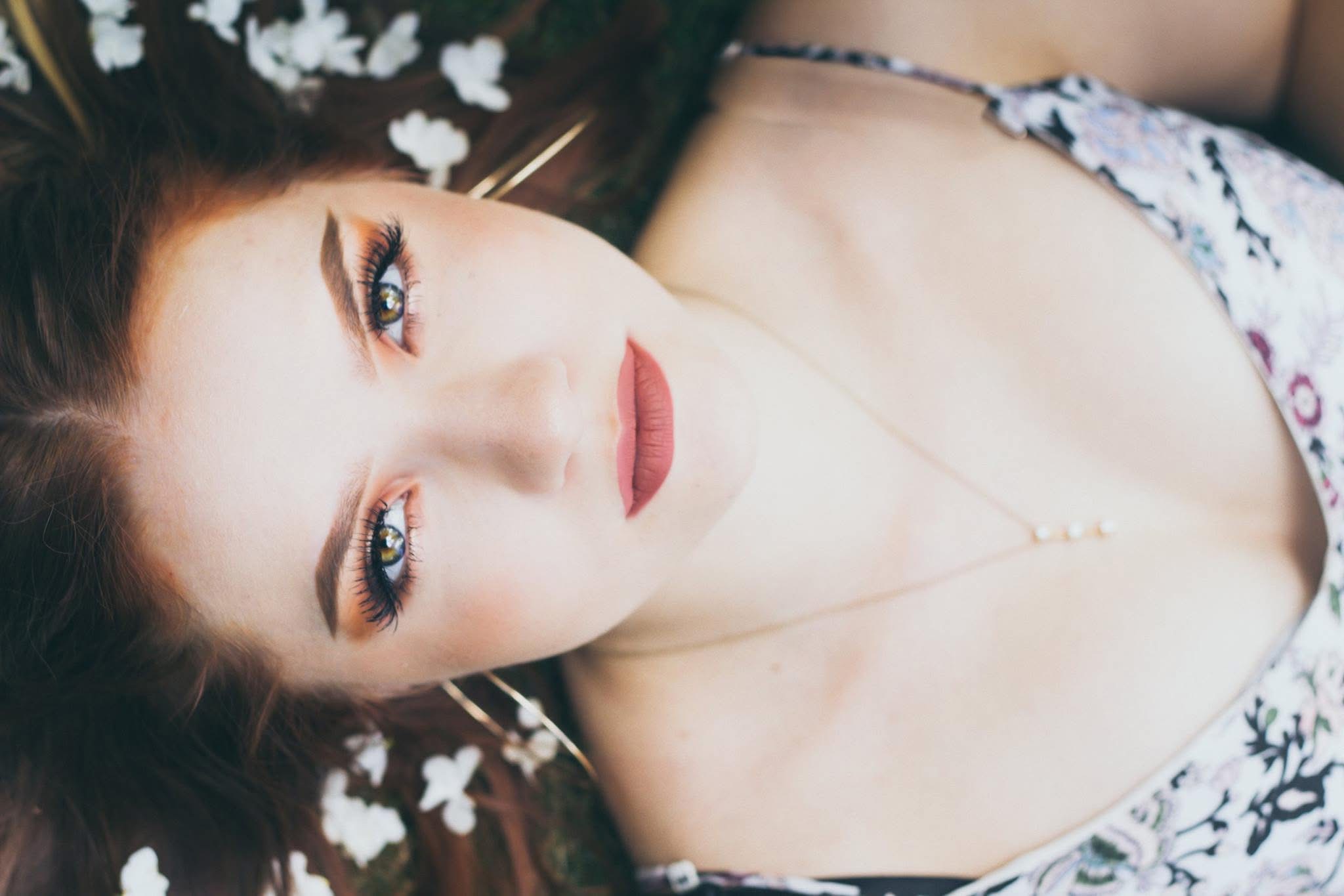 Woman with makeup lying down, looking at the camera, wearing a floral top and a necklace.
