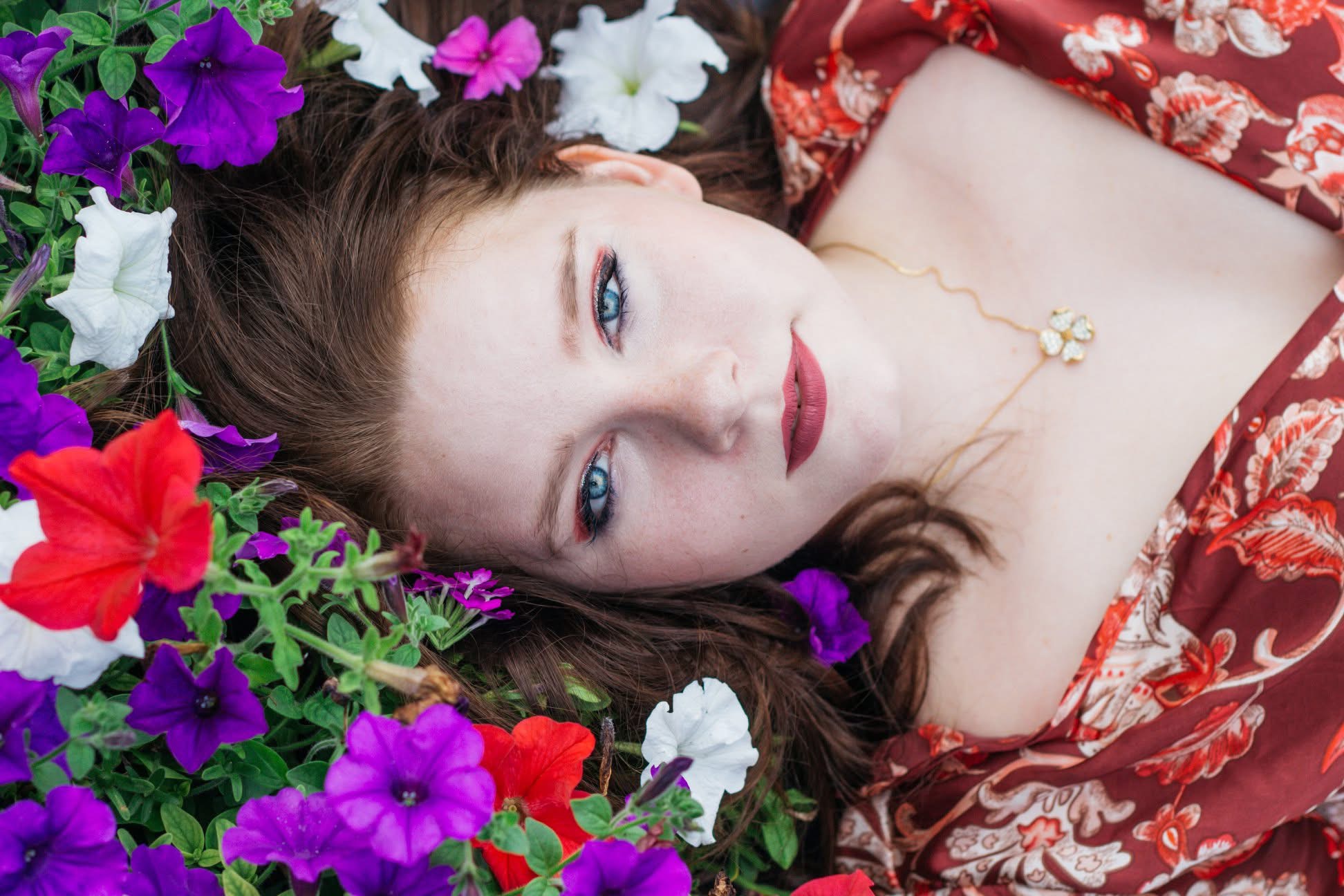 Woman lying among colorful flowers with red, purple, white, and pink blooms, wearing a floral dress and gold necklace.