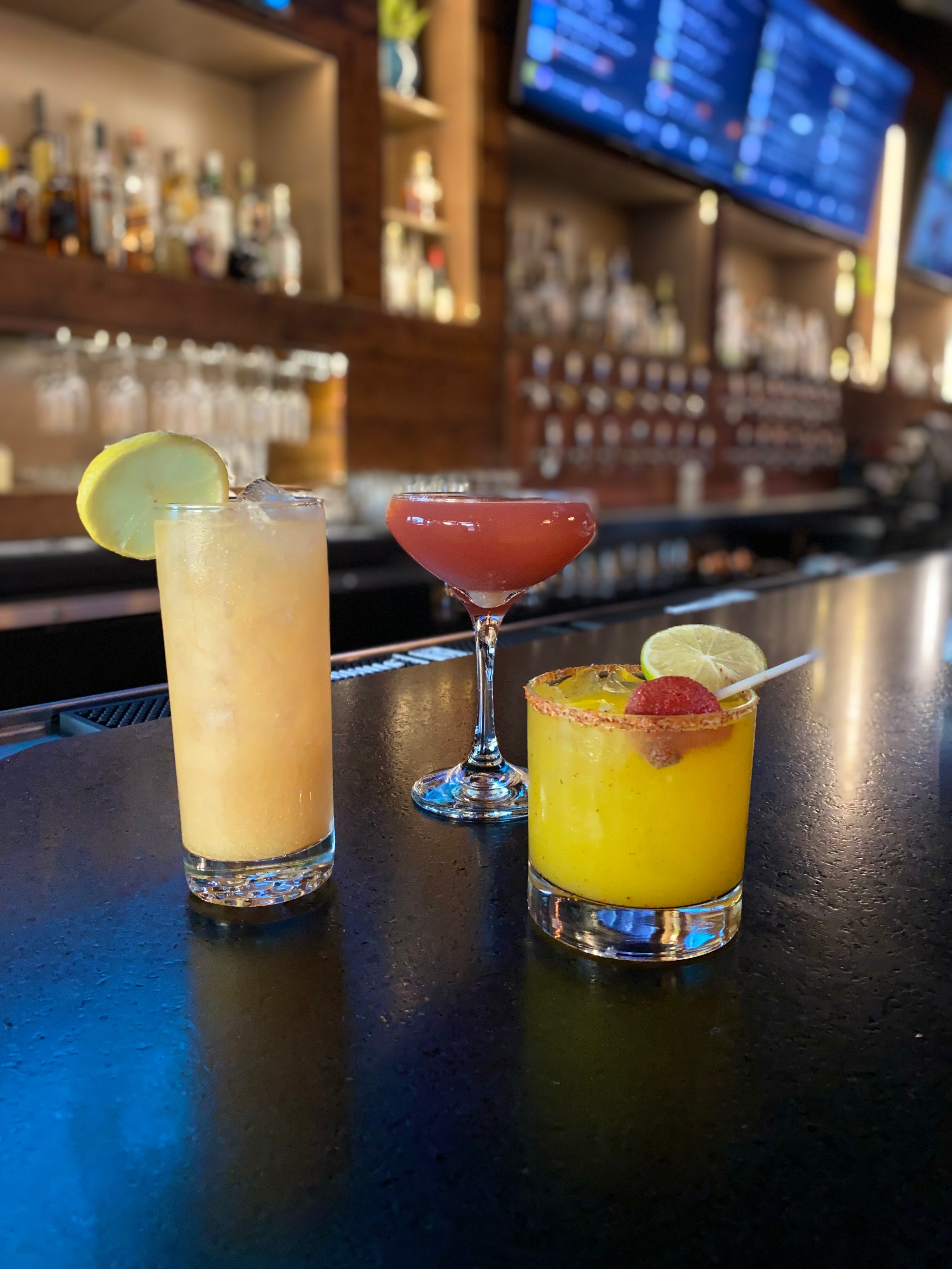 Three colorful cocktails on a bar counter with bottles and a TV screen in the background.