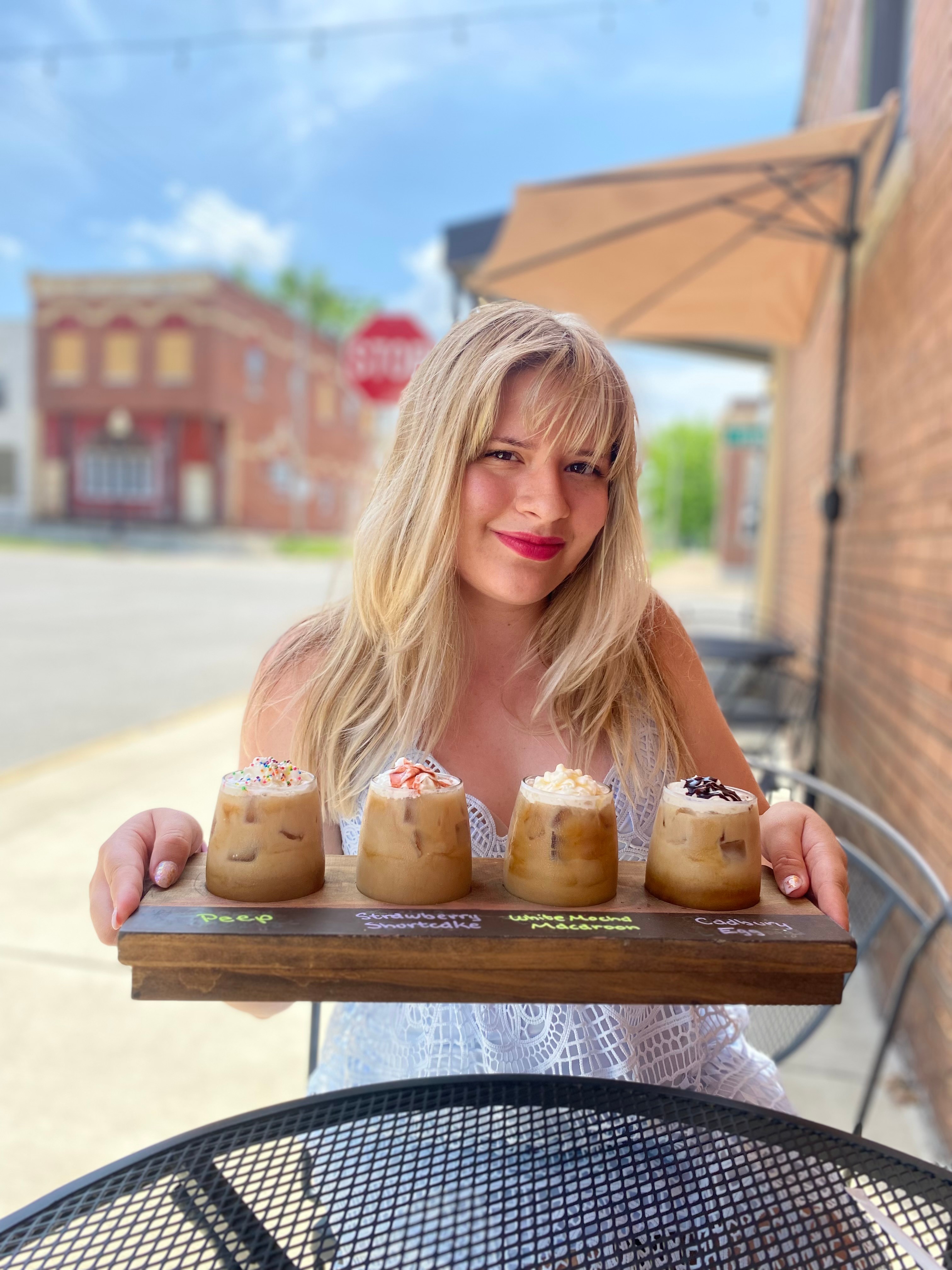 Young woman with blonde hair holding a tray of five cupcakes outdoors, smiling, with a brick building and blue sky in background.