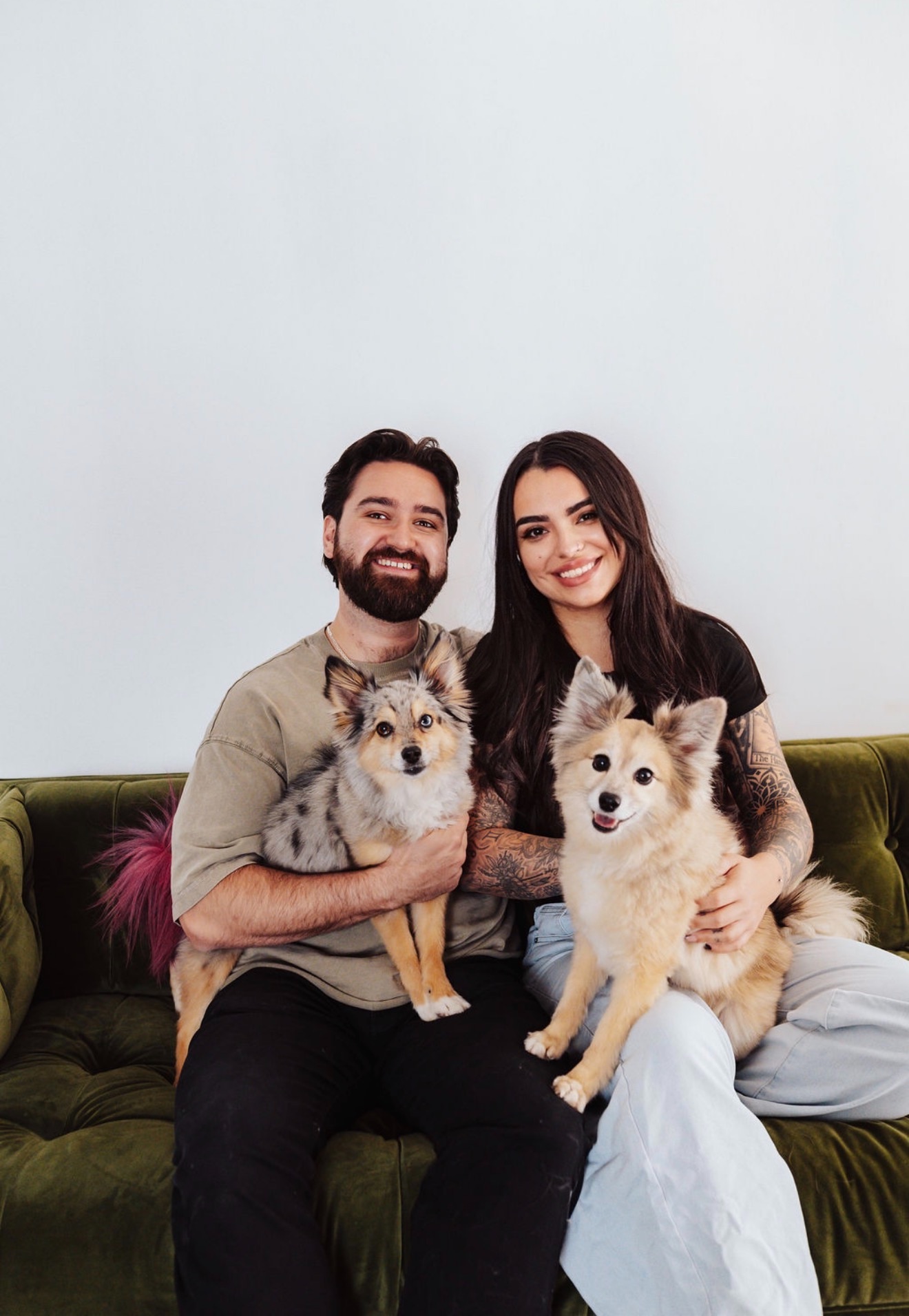 A smiling man and woman sitting on a green sofa, each holding a dog, against a plain light background.