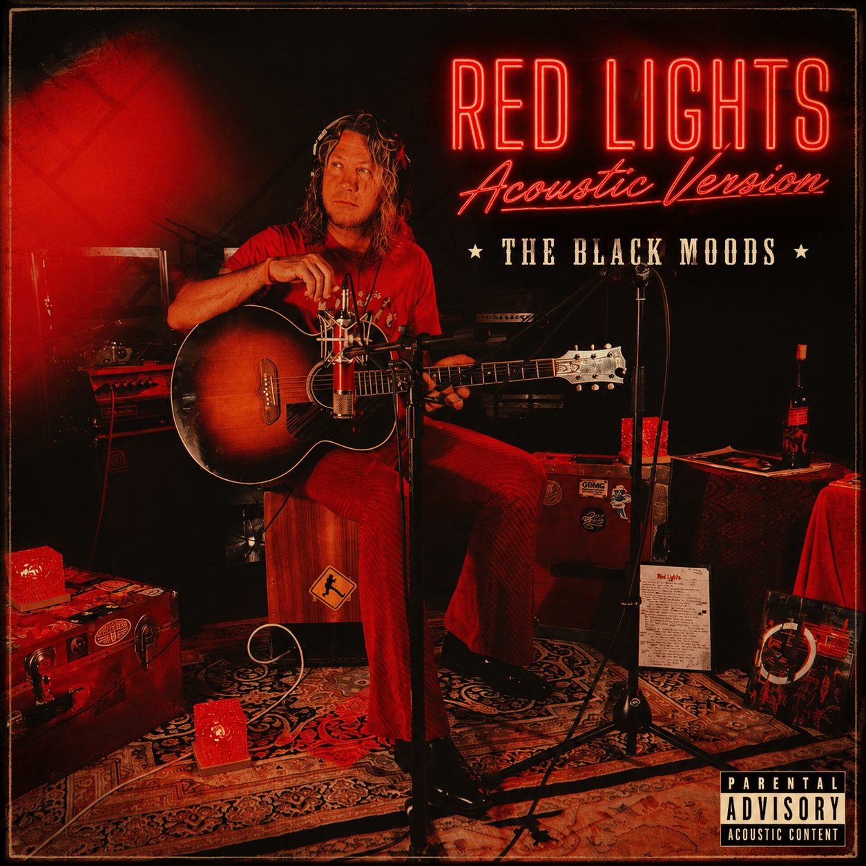 Person sitting with guitar in a music studio, red neon sign reads 'Red Lights Acoustic Version', black background, various equipment around.
