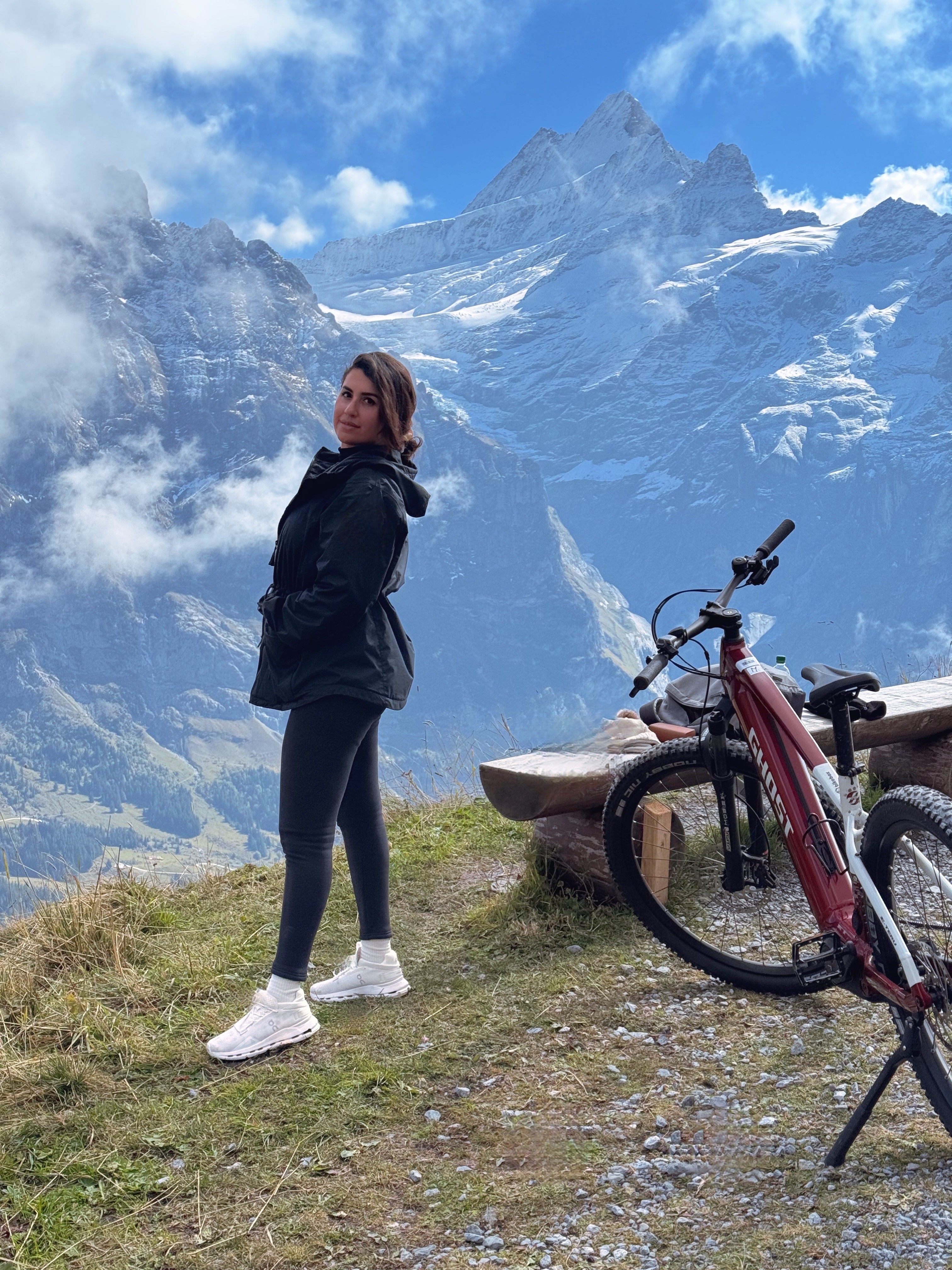 Woman standing outdoors with mountains and clouds in the background, next to a bicycle and a wooden bench.