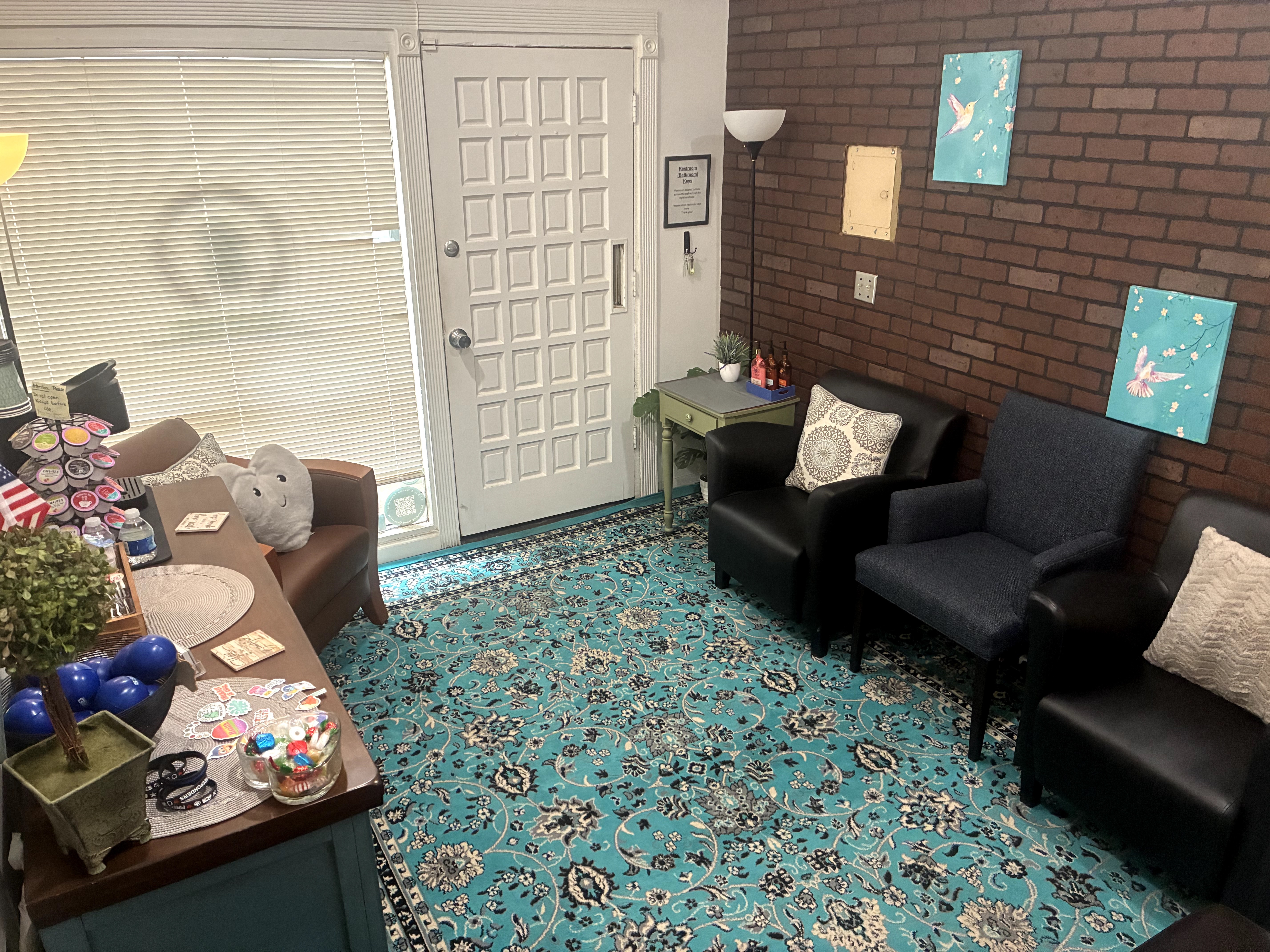 Living room with chairs, a table, and a patterned rug, brick wall with artwork, door, and window with blinds.