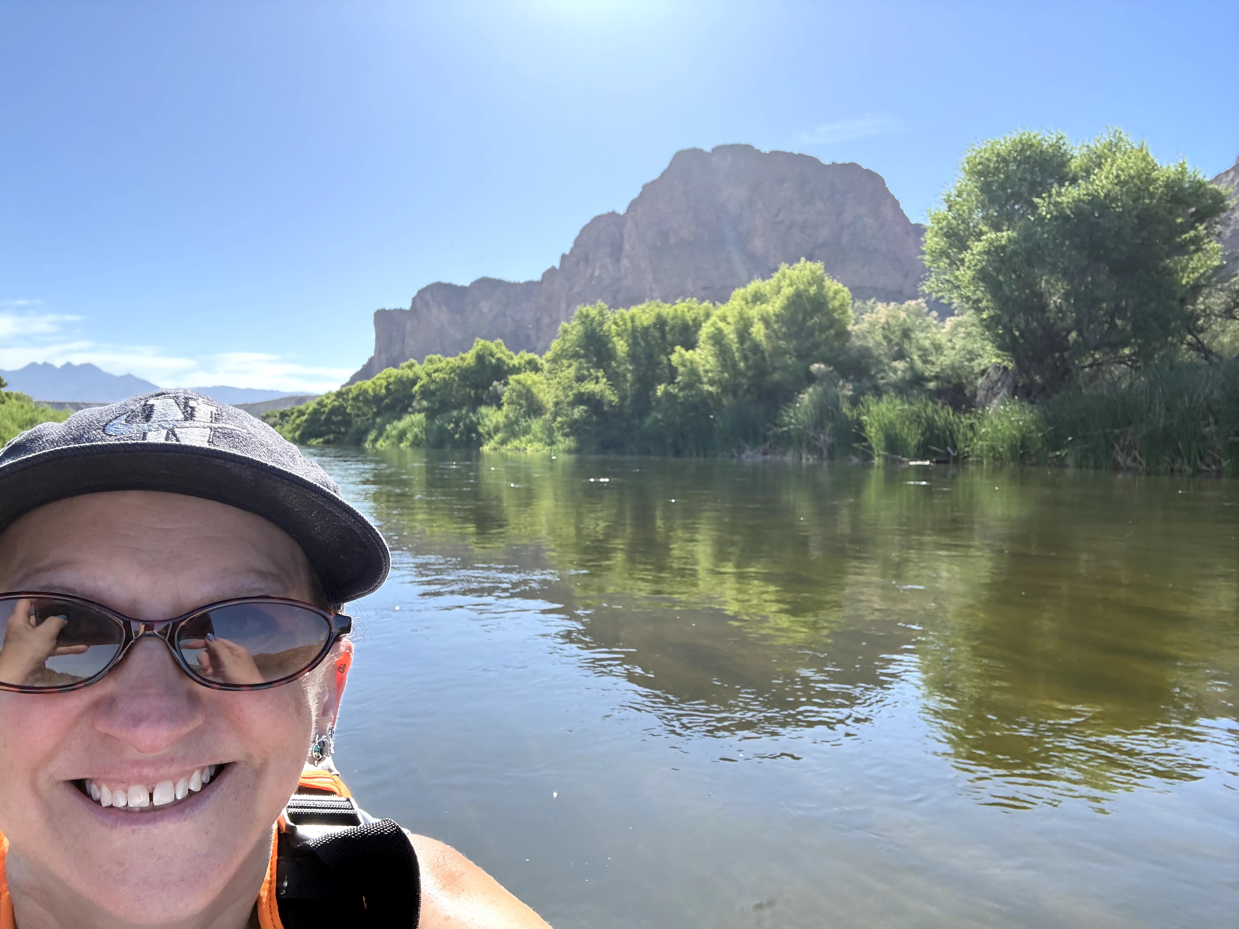 Person wearing sunglasses and a hat smiling near a river with trees and a mountain in the background.