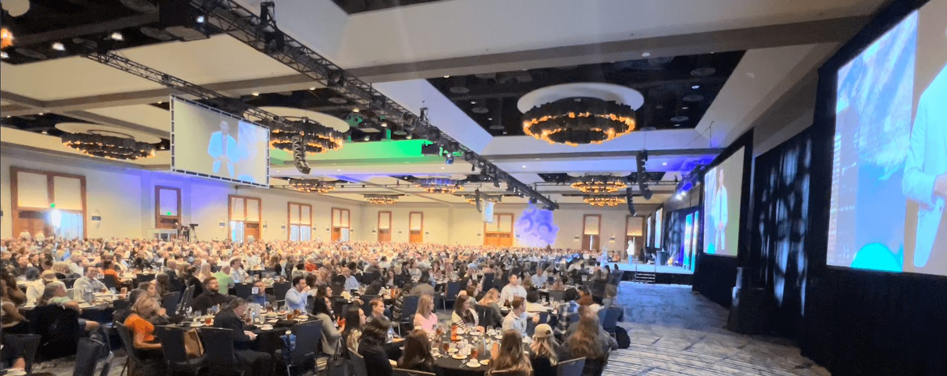Large conference hall filled with seated attendees, multiple screens, and stage lighting, hosting a presentation or event.