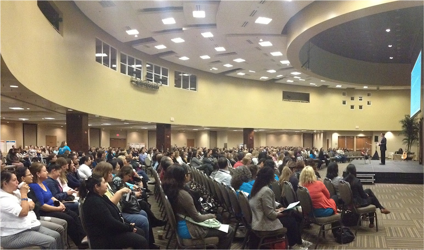 Large audience seated in a spacious auditorium with a stage and curved ceiling, attending a presentation or event.