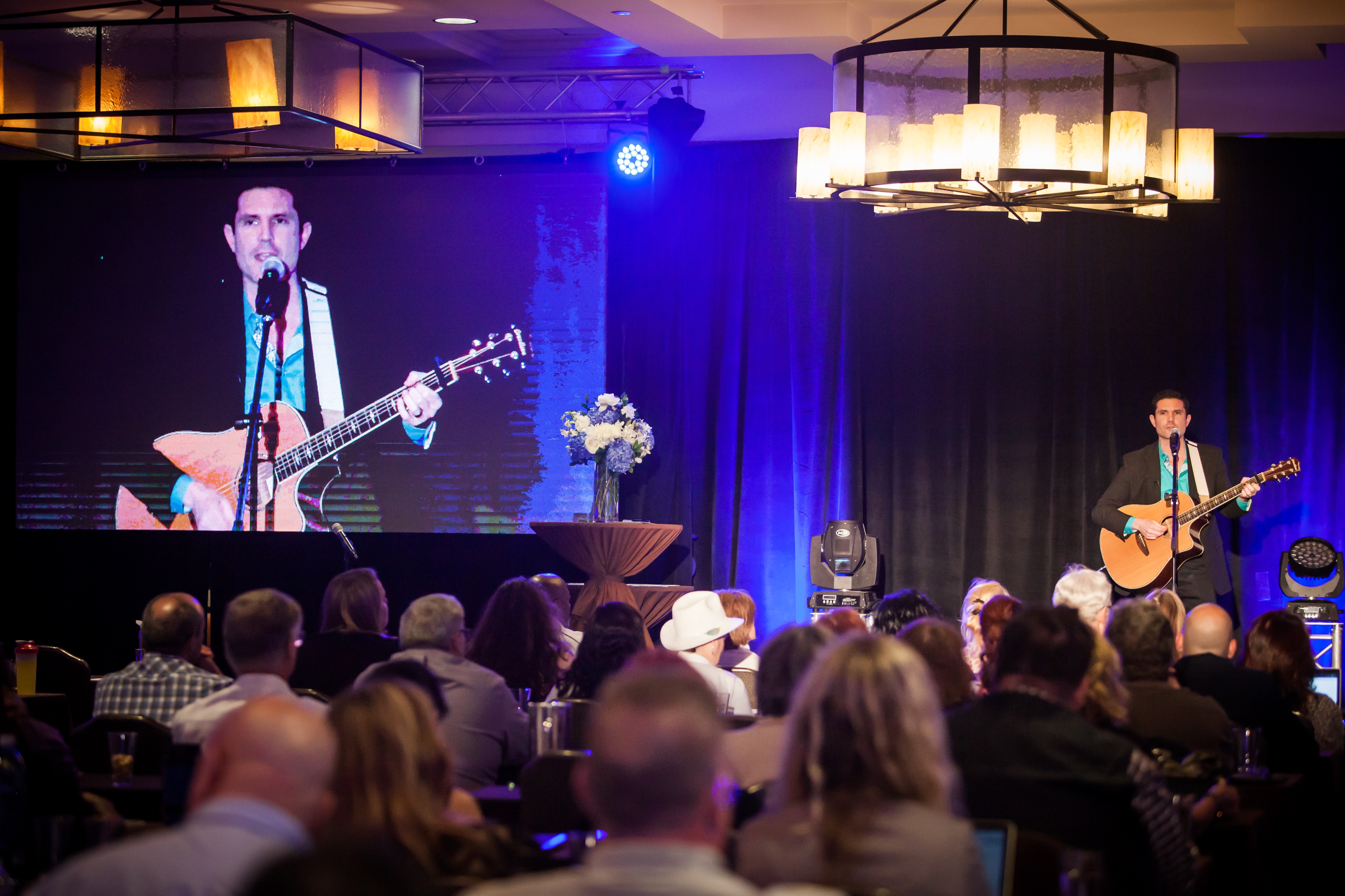 Musicians perform on stage with guitars, large screen shows a close-up of one singer, audience seated watching, stage decorated with flowers.