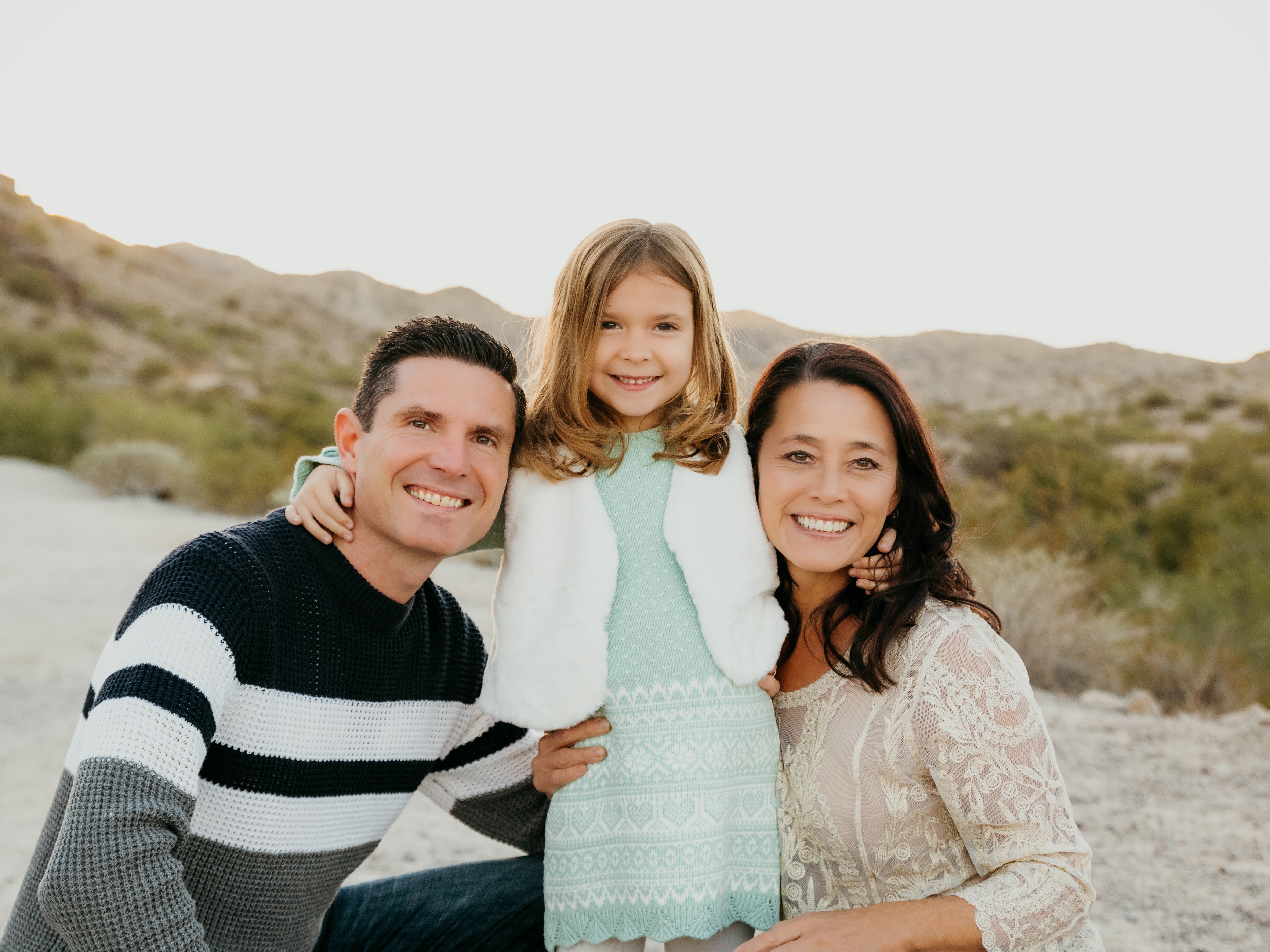 Three people smiling outdoors, a man, a woman, and a girl, with a desert landscape background.