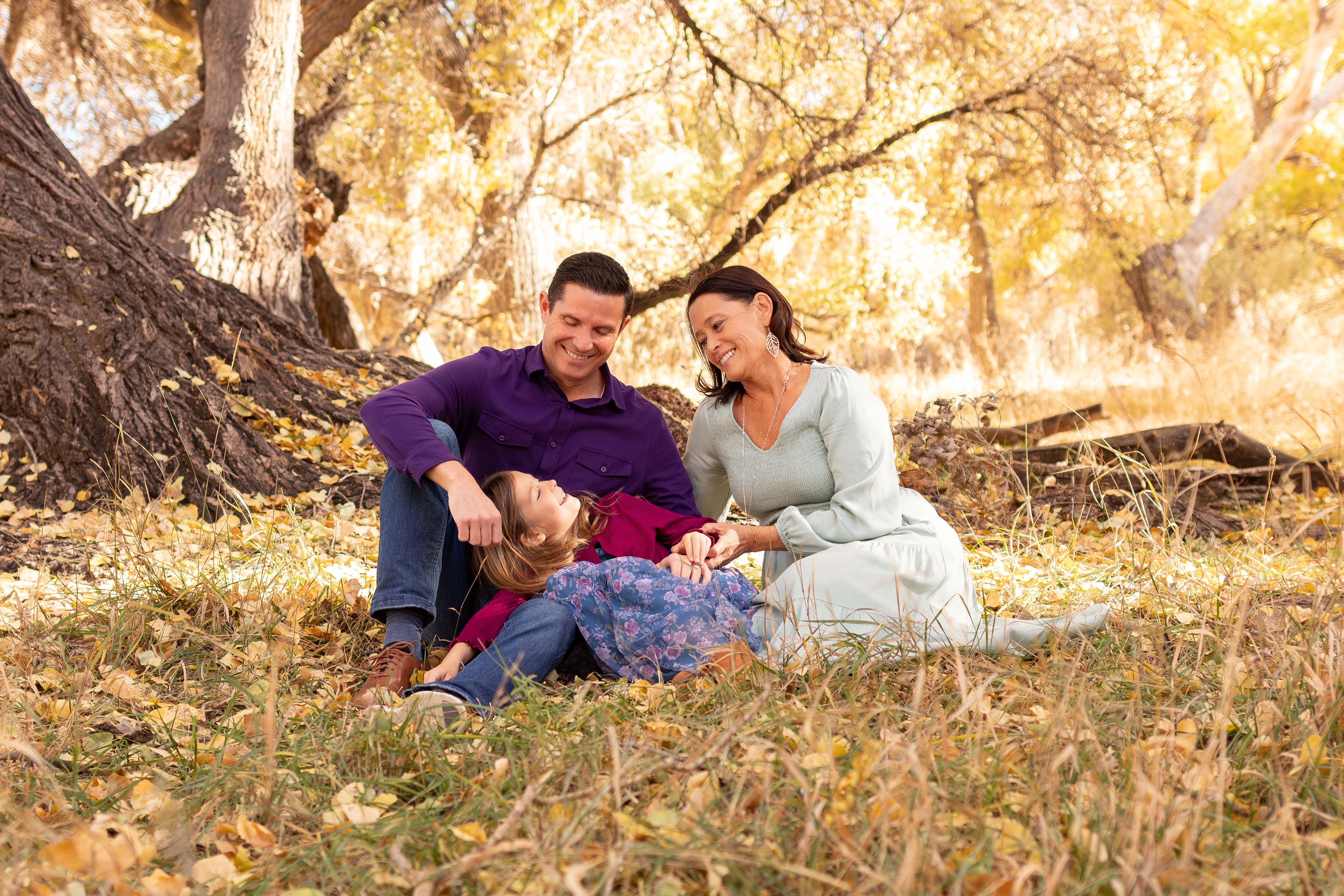 A family sitting on the ground in a park with autumn leaves, trees, and sunlight in the background.