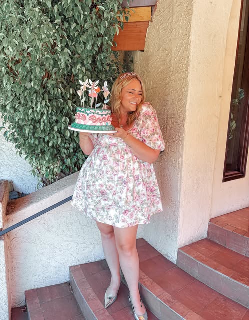Woman with curly hair holding a decorated cake outdoors, standing on steps near a wall and greenery.