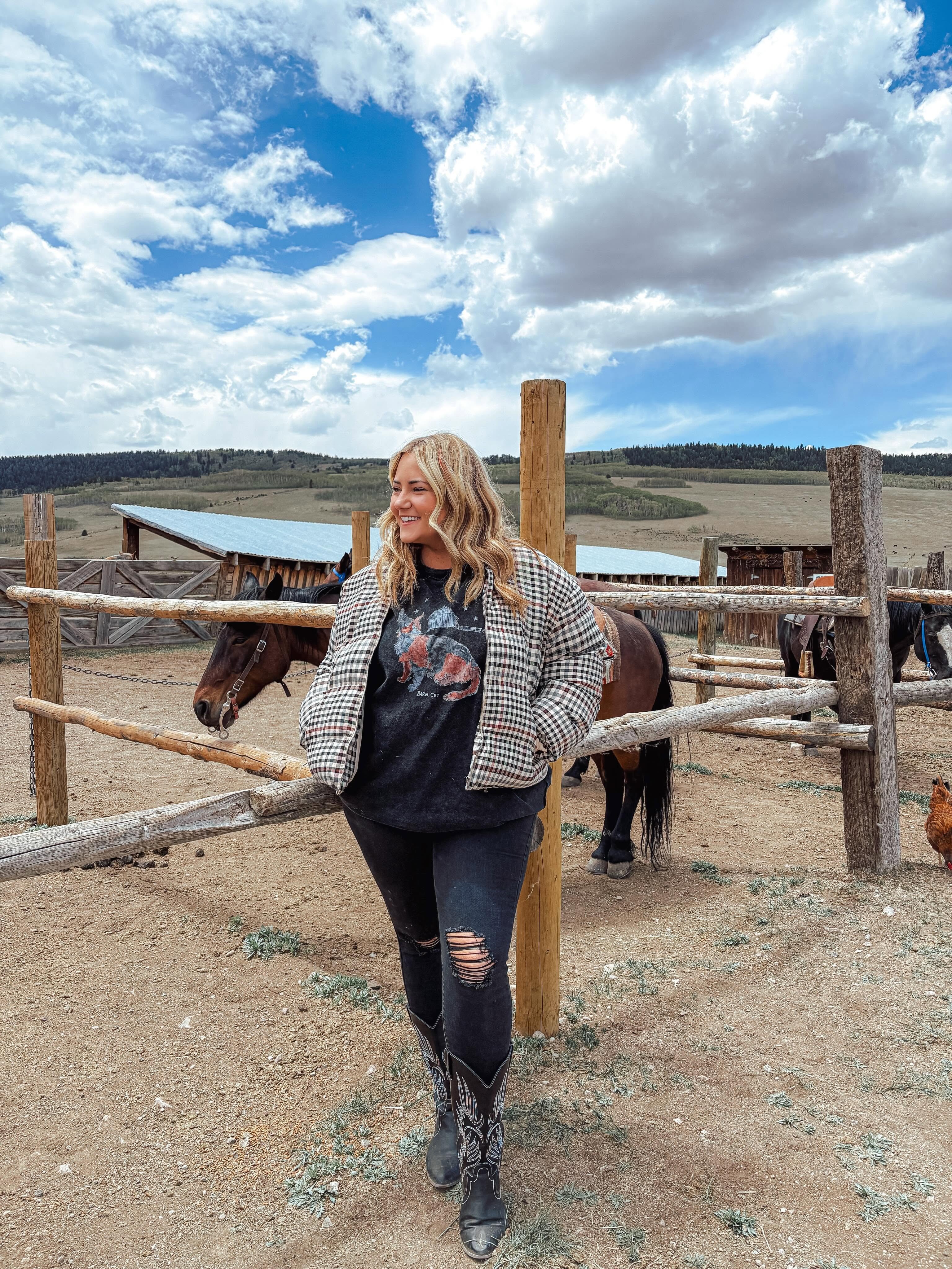 Woman with blonde hair in a checkered jacket and ripped jeans stands near a wooden fence with horses, outdoors under a cloudy sky.