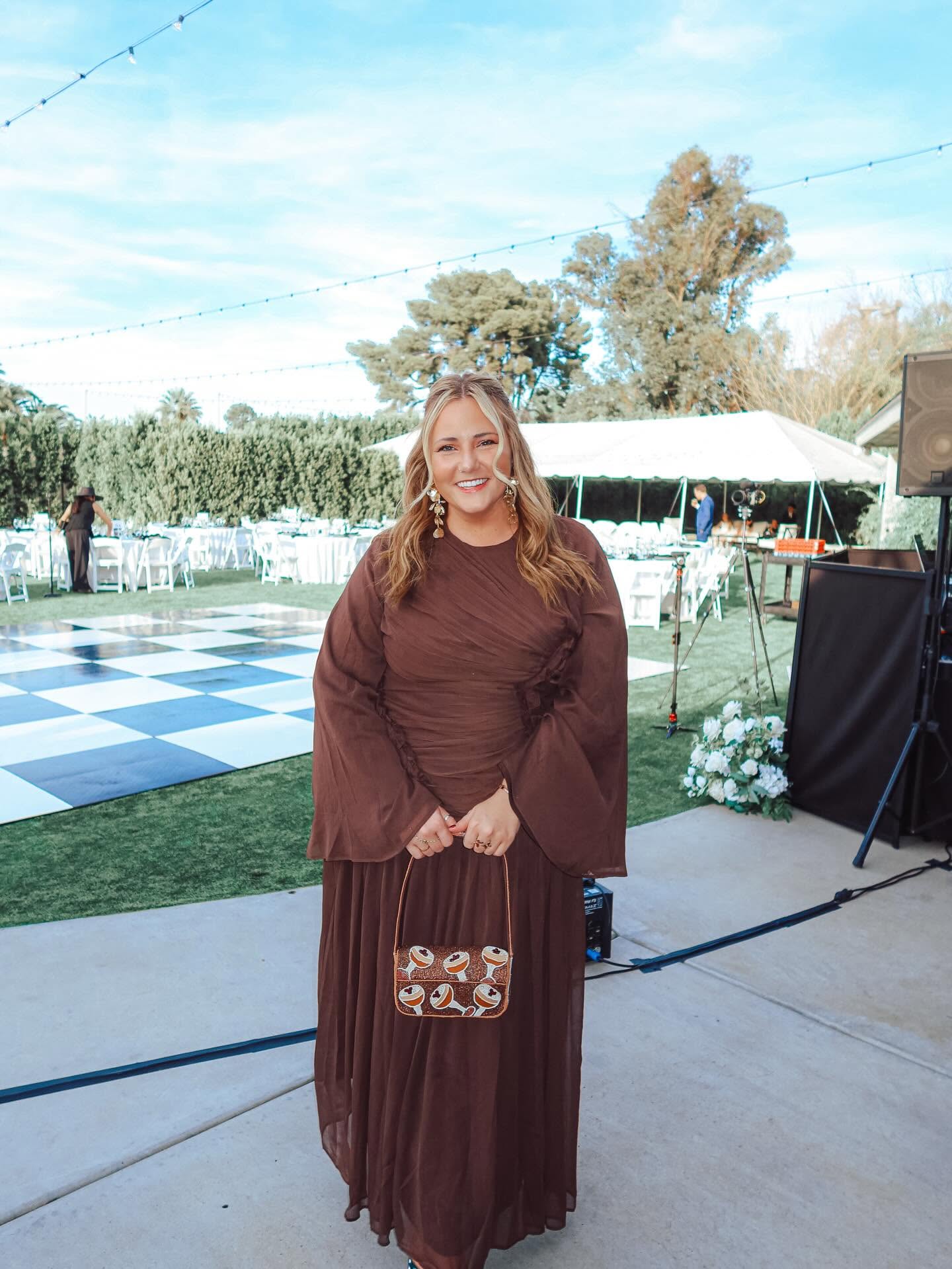 Woman in a long brown dress holding a patterned clutch, smiling outdoors with a tent and trees in the background.