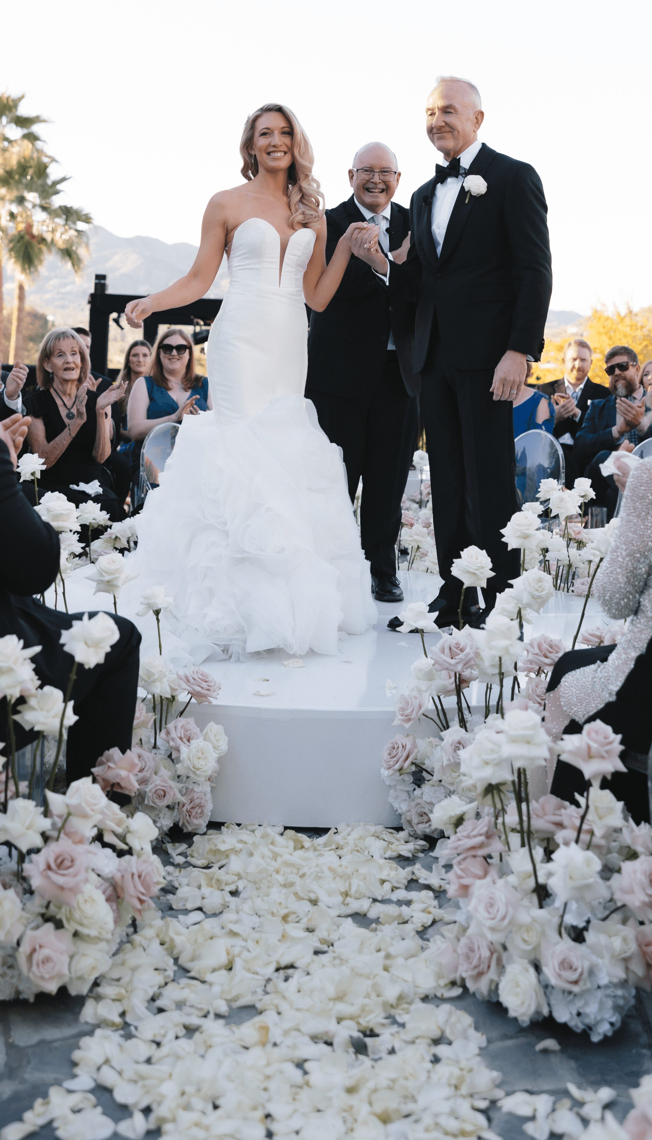 Bride and groom stand together during wedding ceremony, surrounded by seated guests and floral decorations, outdoors with mountains in background.