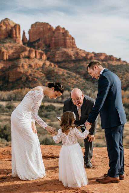 A family of four holding hands in a desert landscape with red rock formations, dressed in formal attire, smiling.