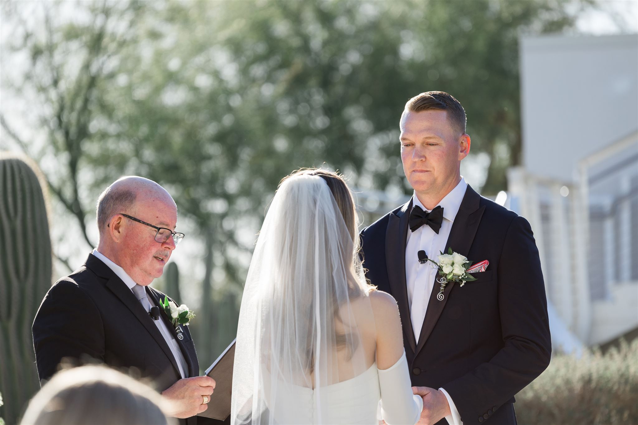 A wedding ceremony outdoors with a bride, groom, and officiant, all dressed formally, with trees and a white structure in background.