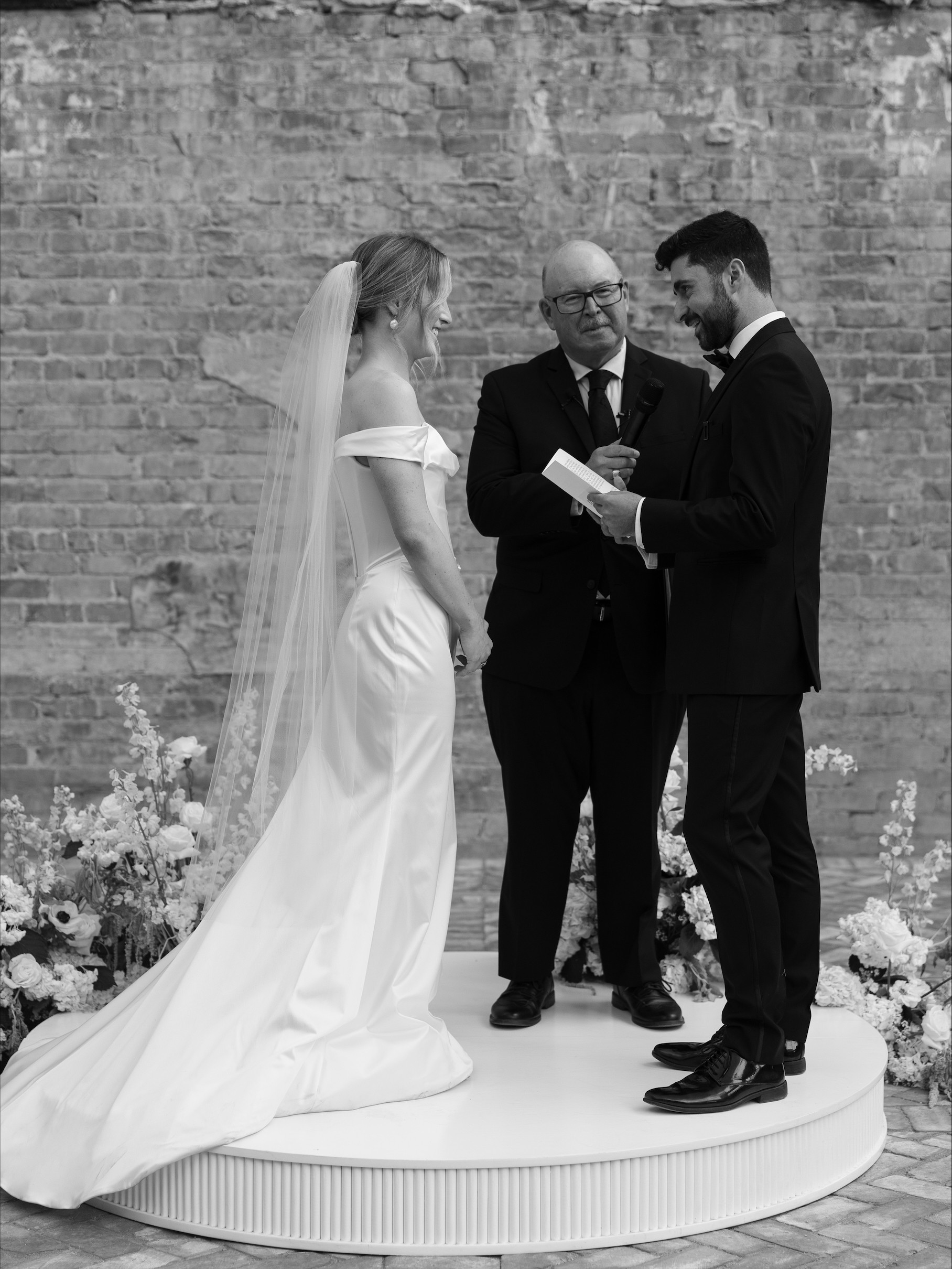 Bride and groom stand on a platform with officiant, reading vows, against a brick wall, in black and white.