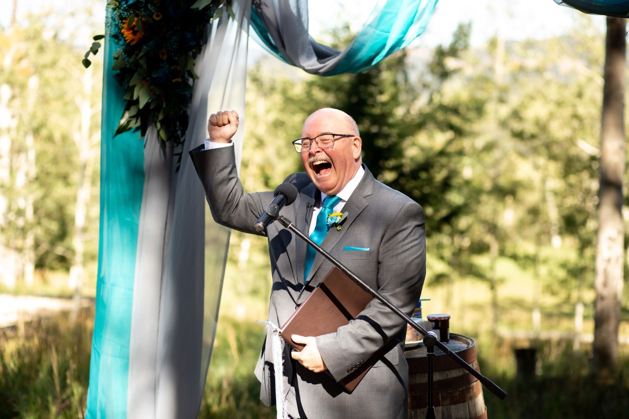 Man in suit celebrating with raised fist at outdoor event, holding a folder, with trees and decorations in background.