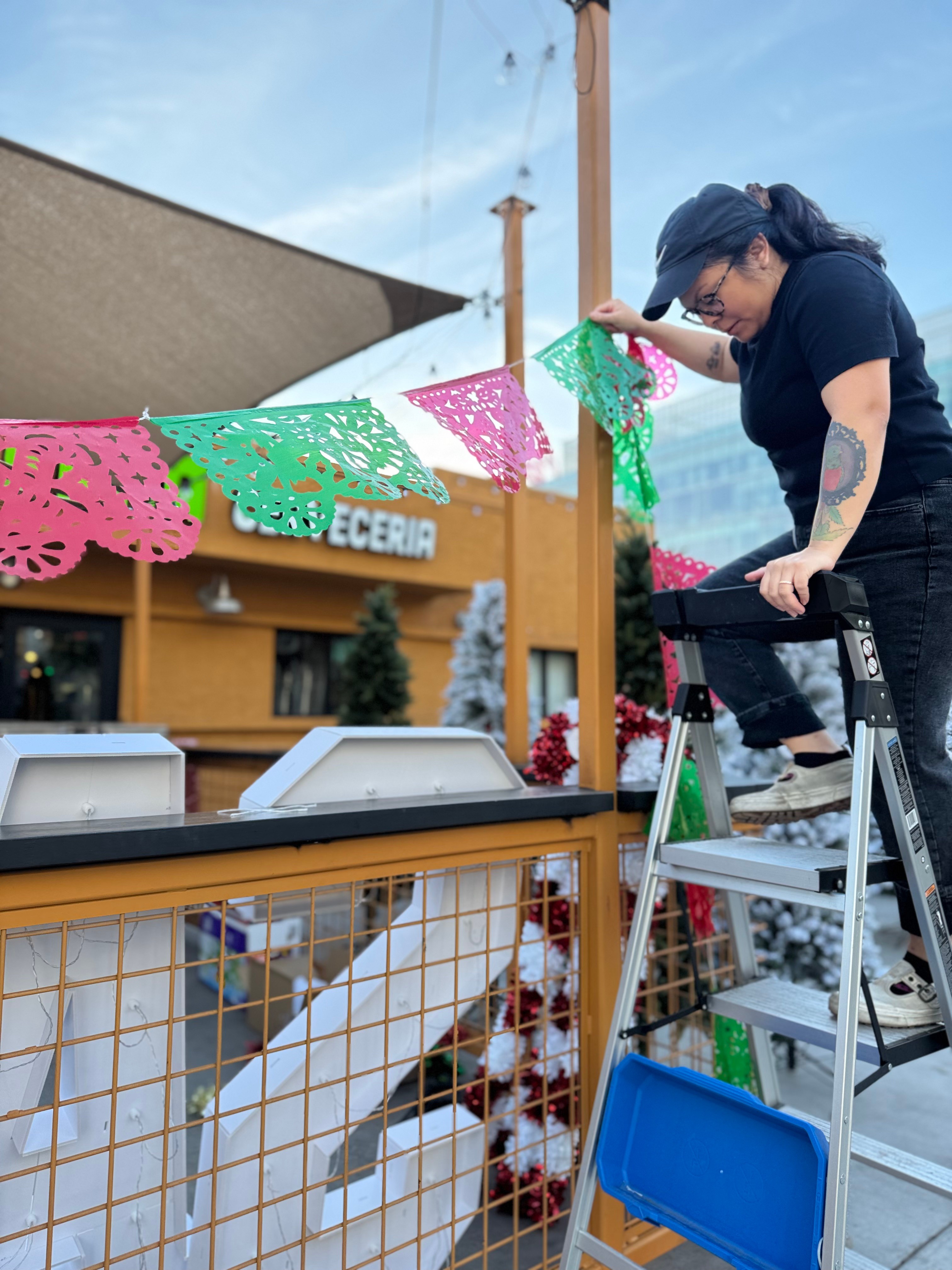 Woman on ladder hanging colorful paper banners outdoors with a building and blue sky in background.