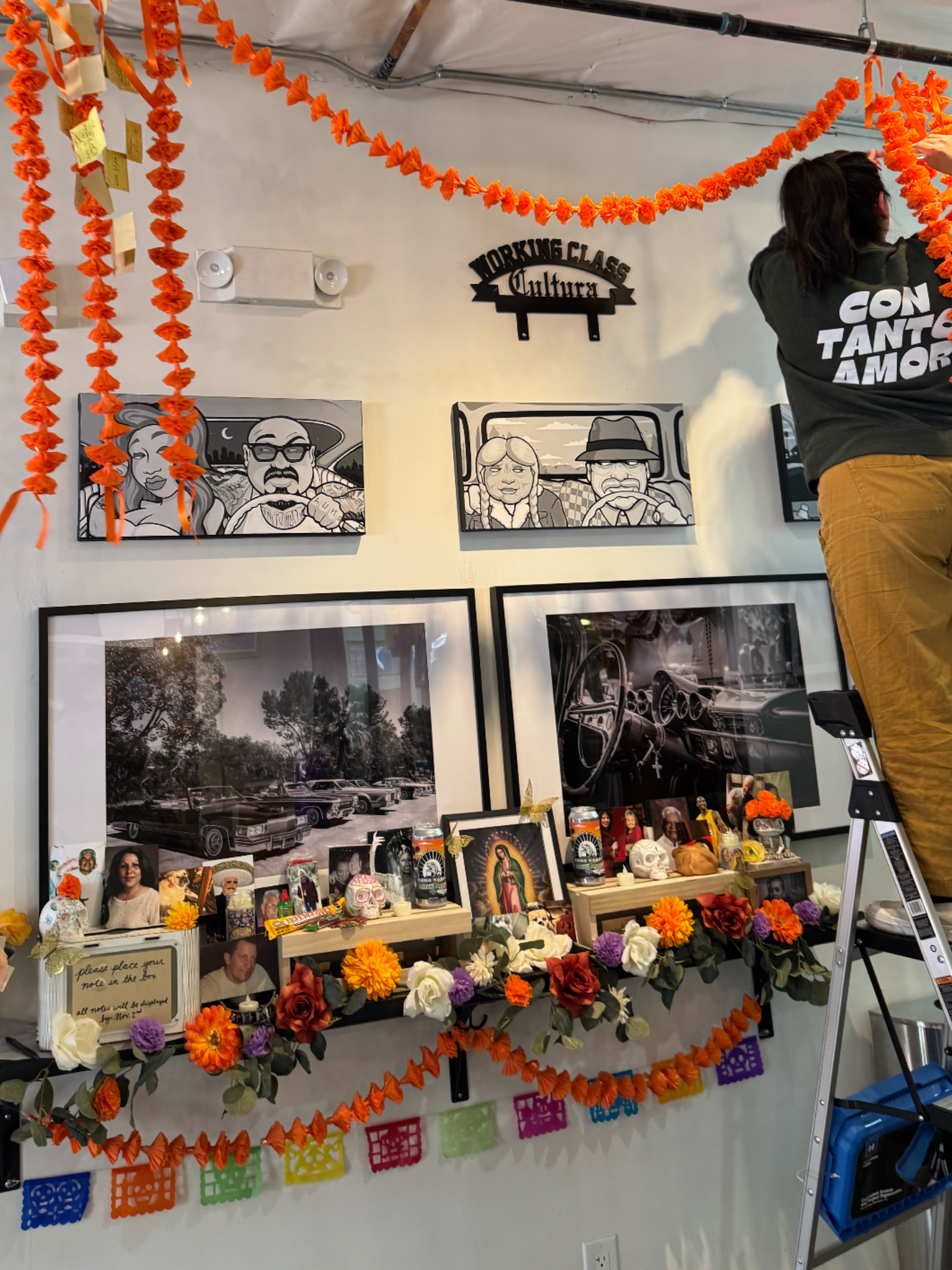 Display of framed photos and artwork with orange paper flowers and colorful banners, part of a festive celebration setup.