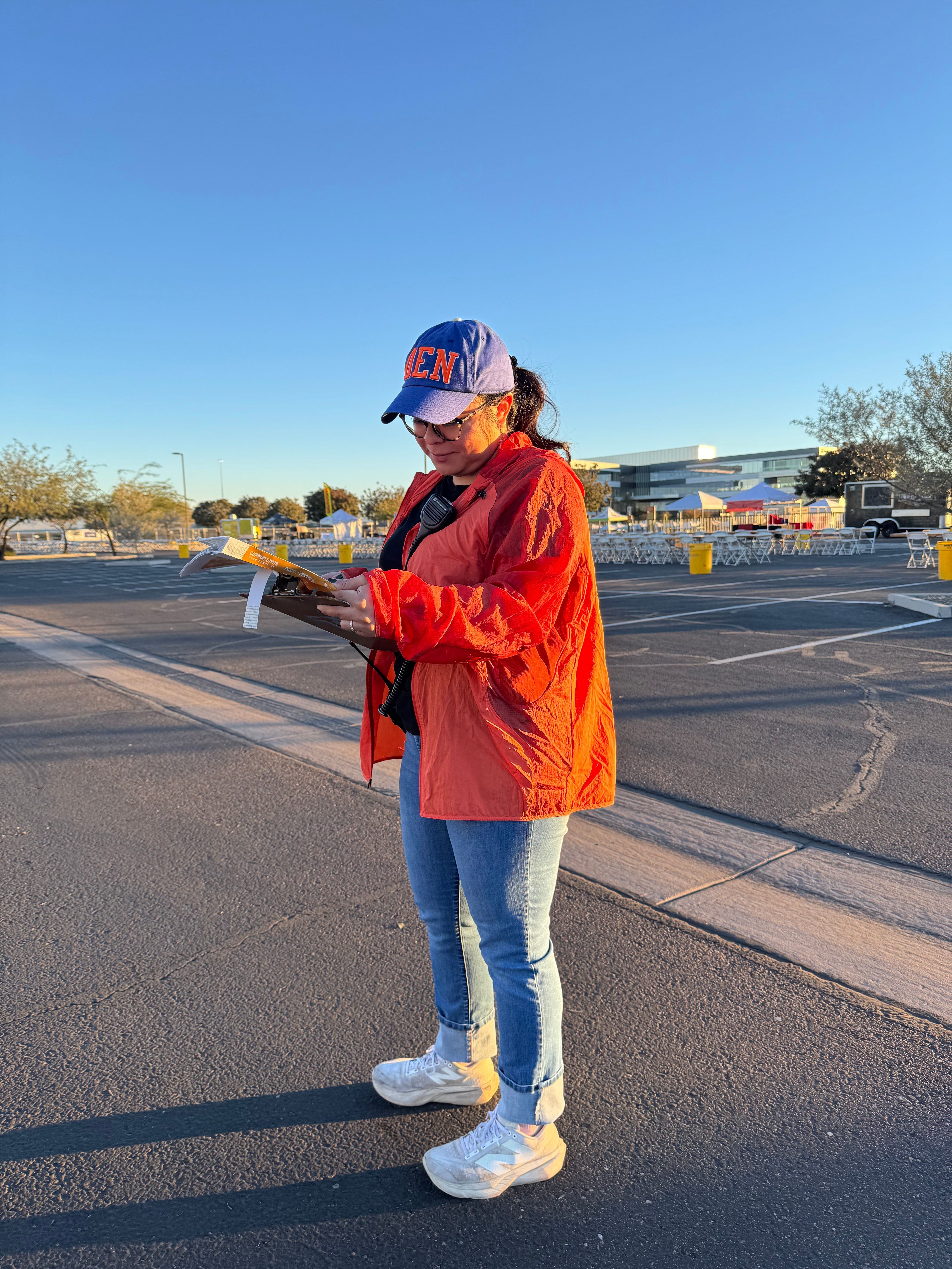 Person in orange jacket and jeans standing on street, holding a phone, with clear sky and buildings in background.