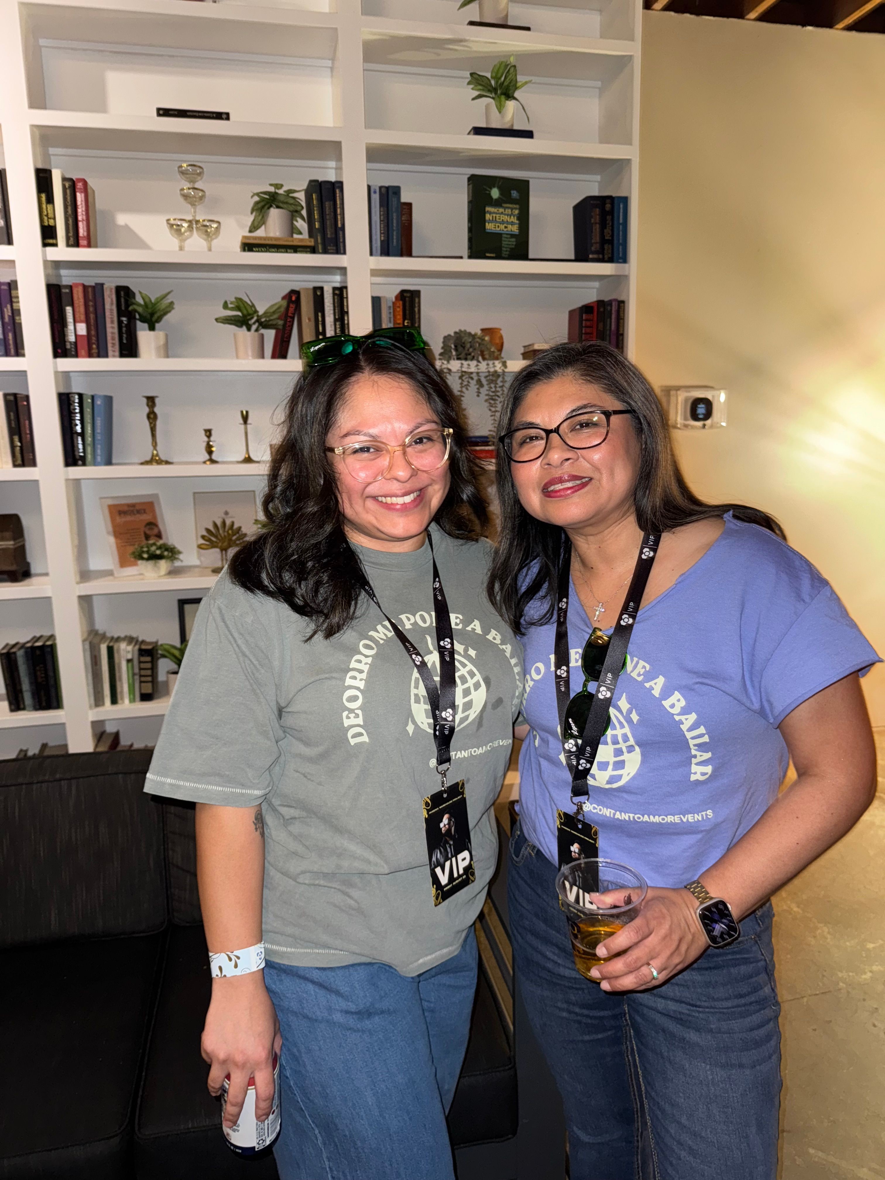 Two women standing in front of a white bookshelf with books and plants, smiling at the camera.