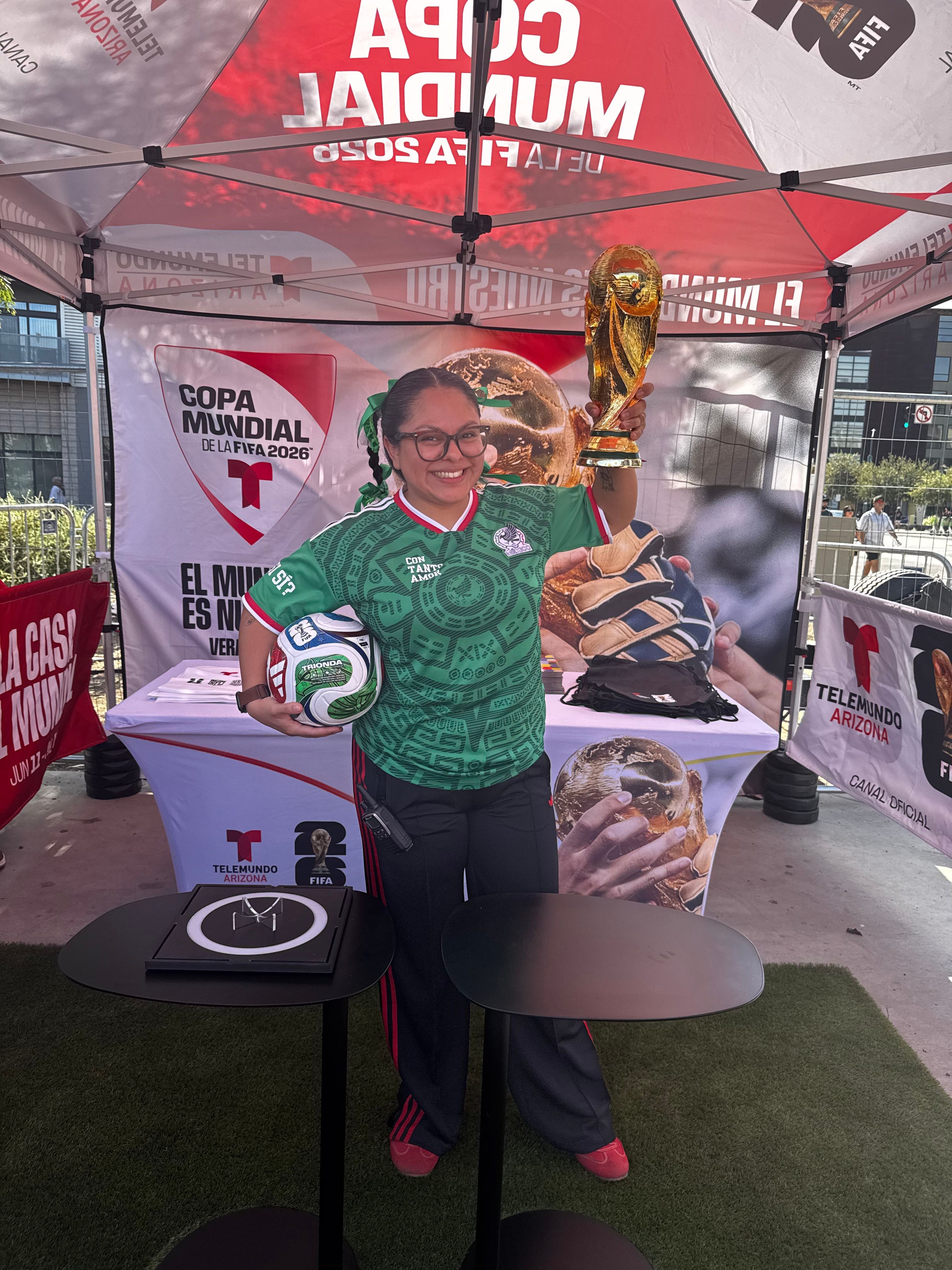 Person holding a trophy at an outdoor event with banners and a table, wearing glasses and a green shirt.