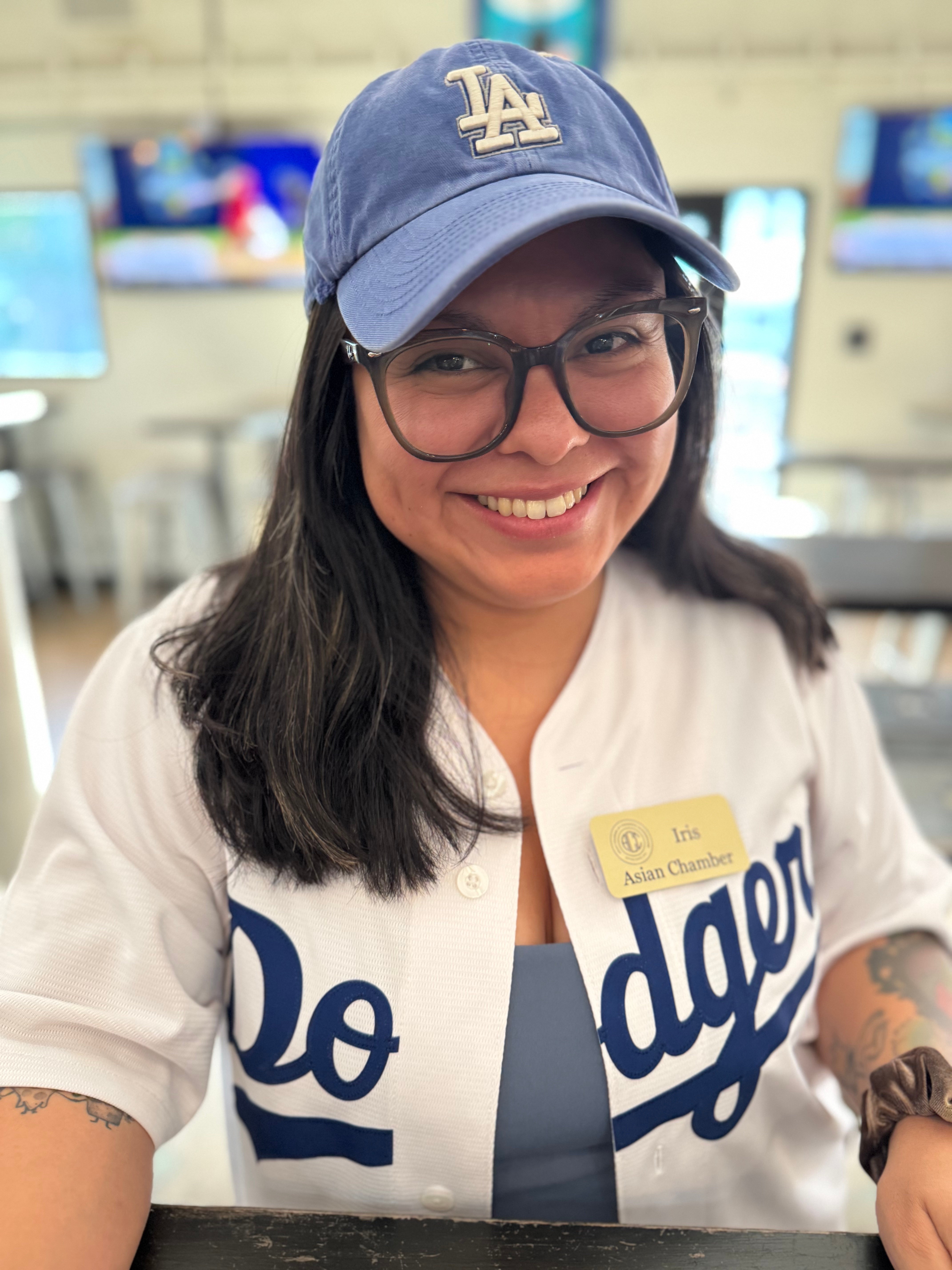 Smiling woman wearing glasses, a Los Angeles Dodgers cap, and a Dodgers jersey in a store, with TV screens in background.