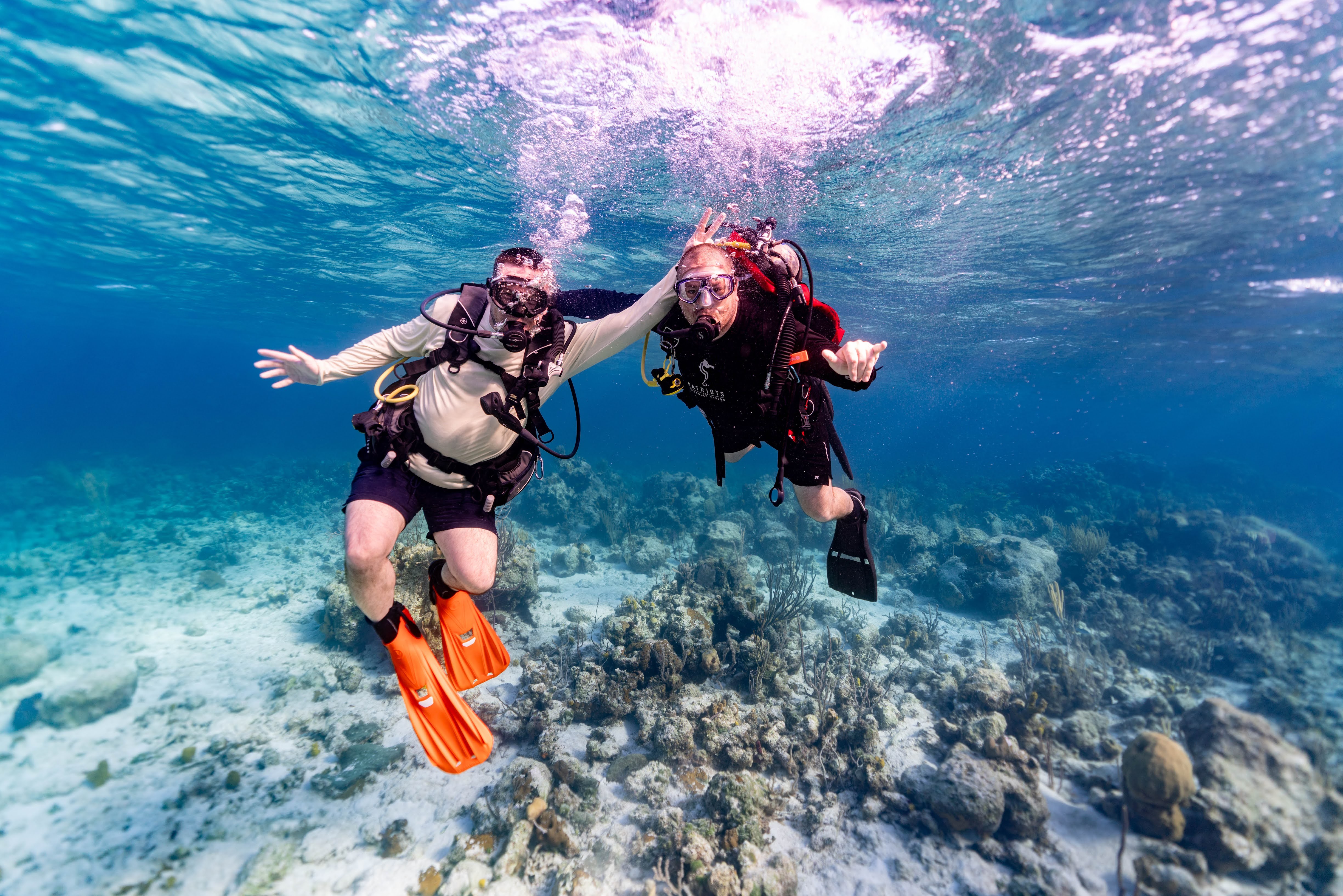 Two people scuba diving underwater over a coral reef, holding hands, with bubbles and clear water above.