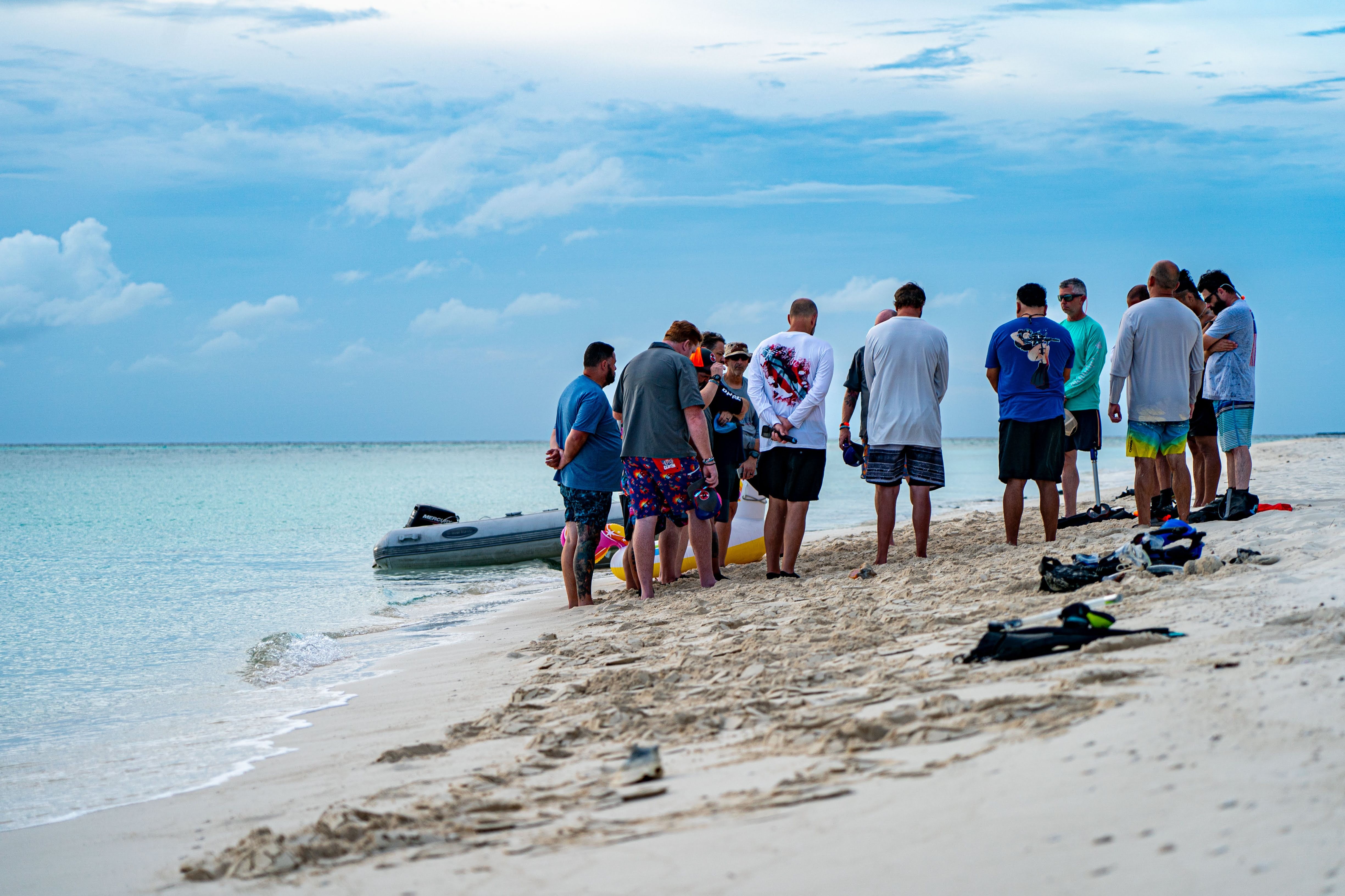 Group of people standing on a sandy beach near the water, some looking at a boat, under a partly cloudy sky.