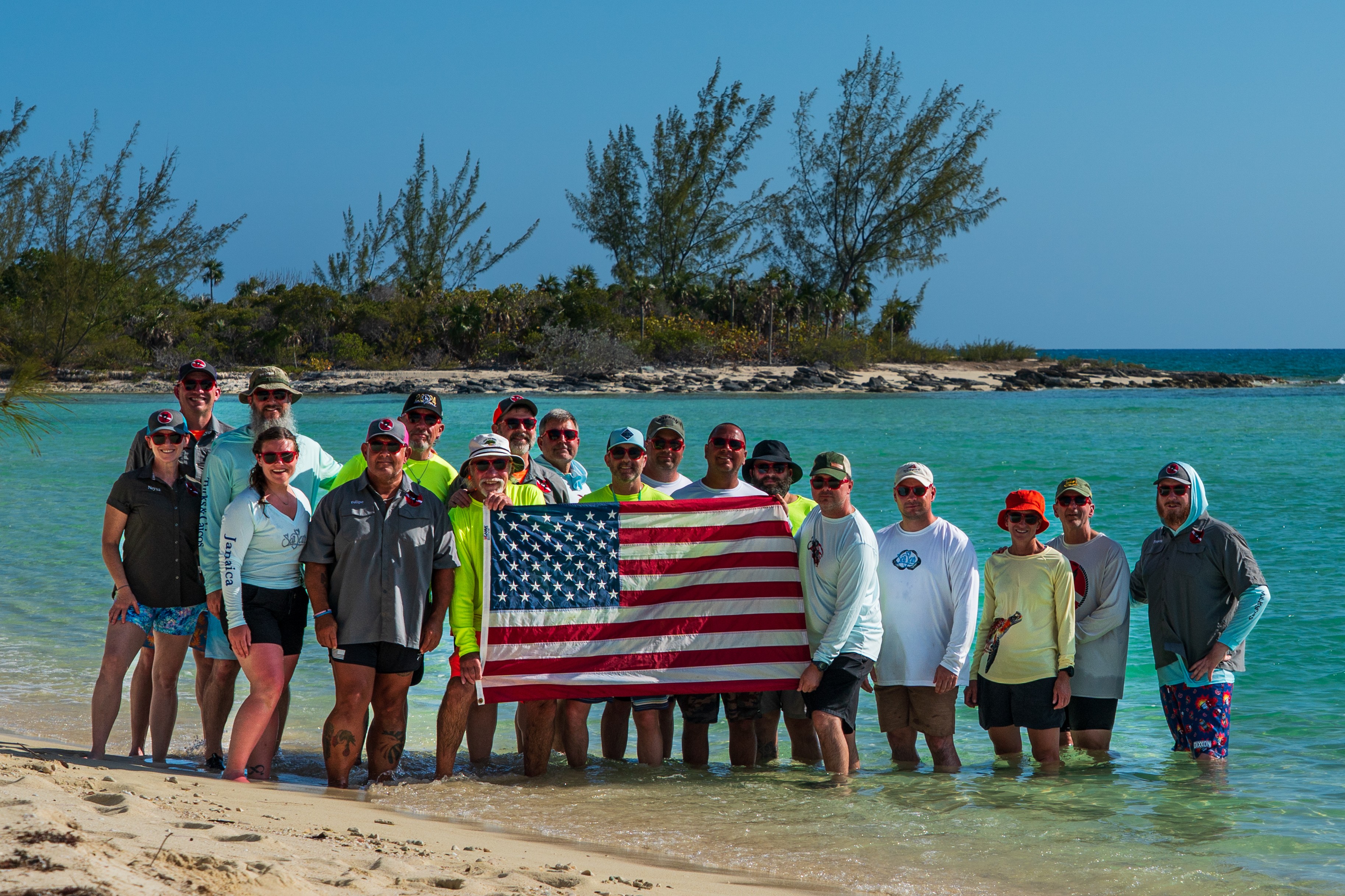 Group of people holding an American flag at the beach with trees and ocean in background.