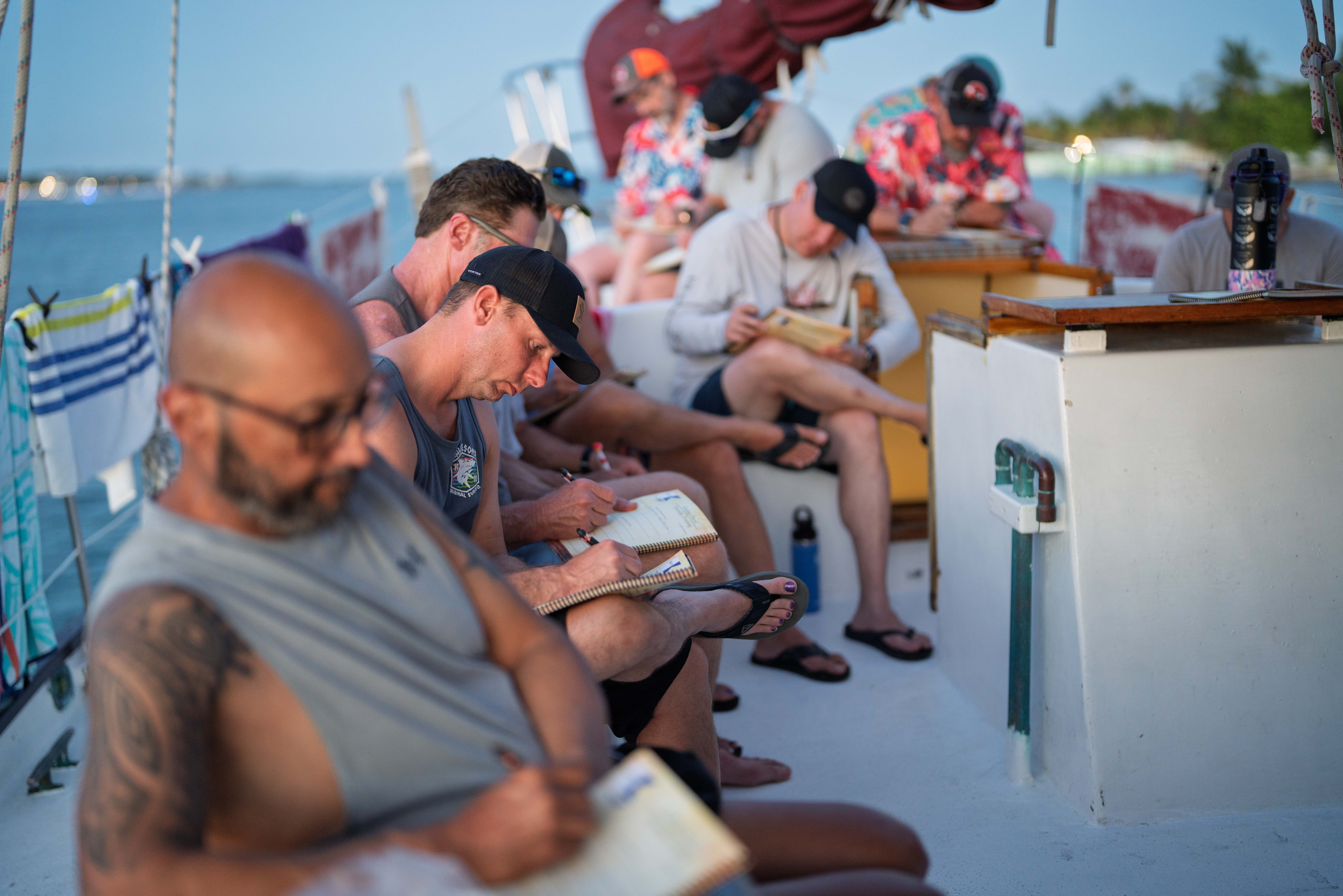 People sitting on a boat deck, some writing or reading, with a body of water and boats in the background.