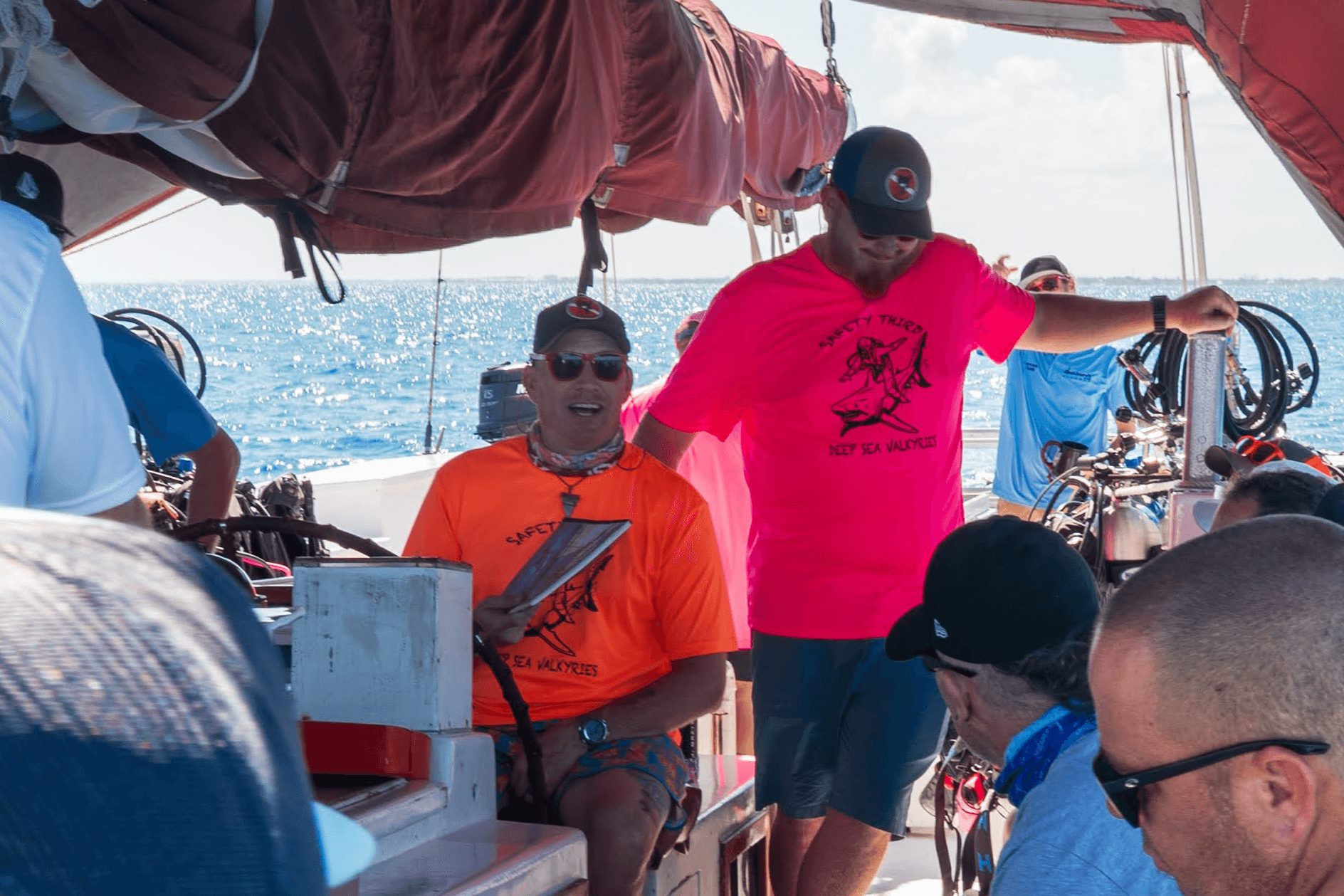 Group of people on a boat, some wearing sunglasses, with water and sky in the background.