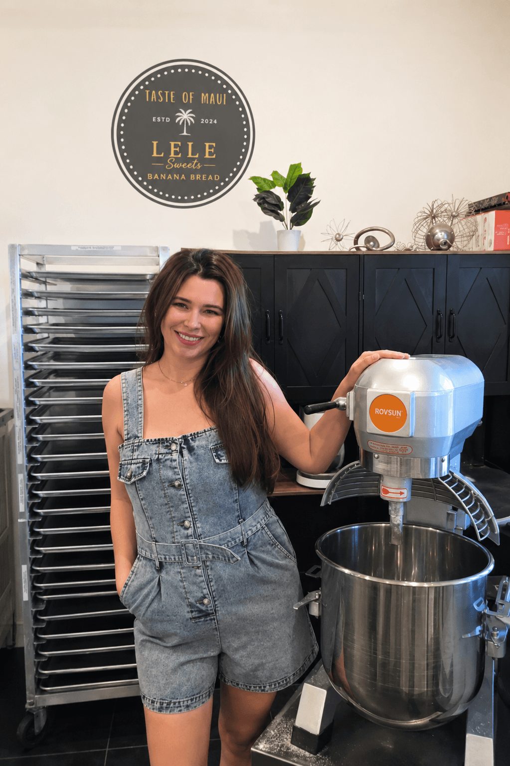 Smiling woman with long hair in denim overalls stands in a kitchen, holding a mixer over a large bowl.