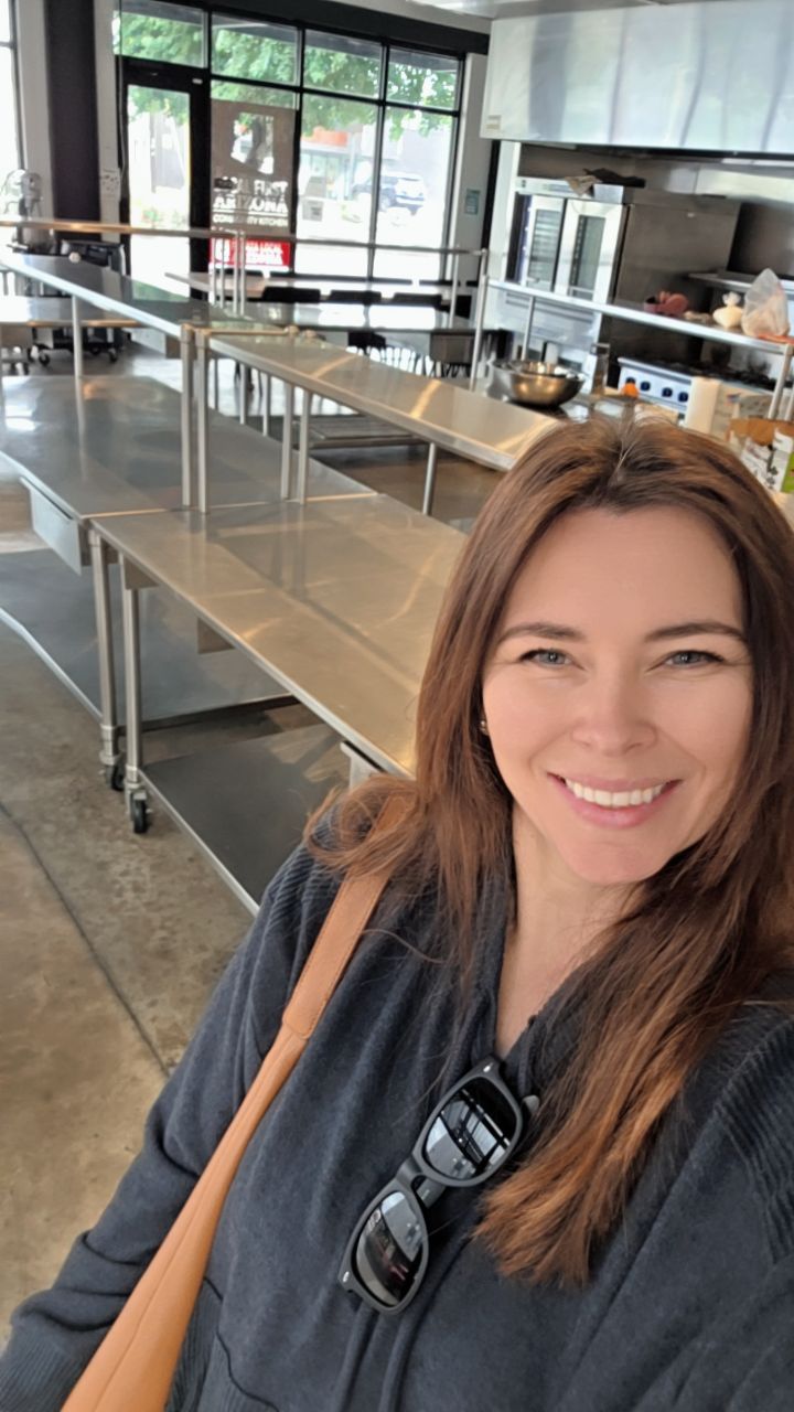 Smiling woman with brown hair in a black top, in a modern kitchen or cafeteria setting with stainless steel counters and large windows.