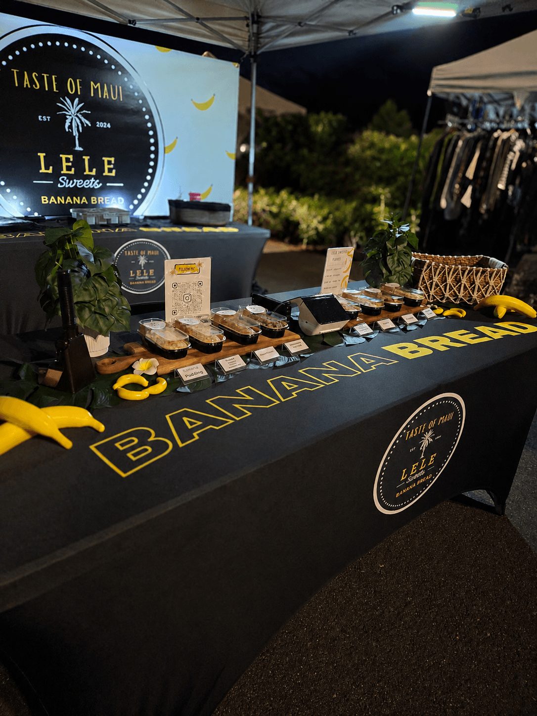 Night market stall with banana bread and banana-related products, illuminated by a light, with a sign reading 'Taste of Maui' and 'Lele Banana Bread'.
