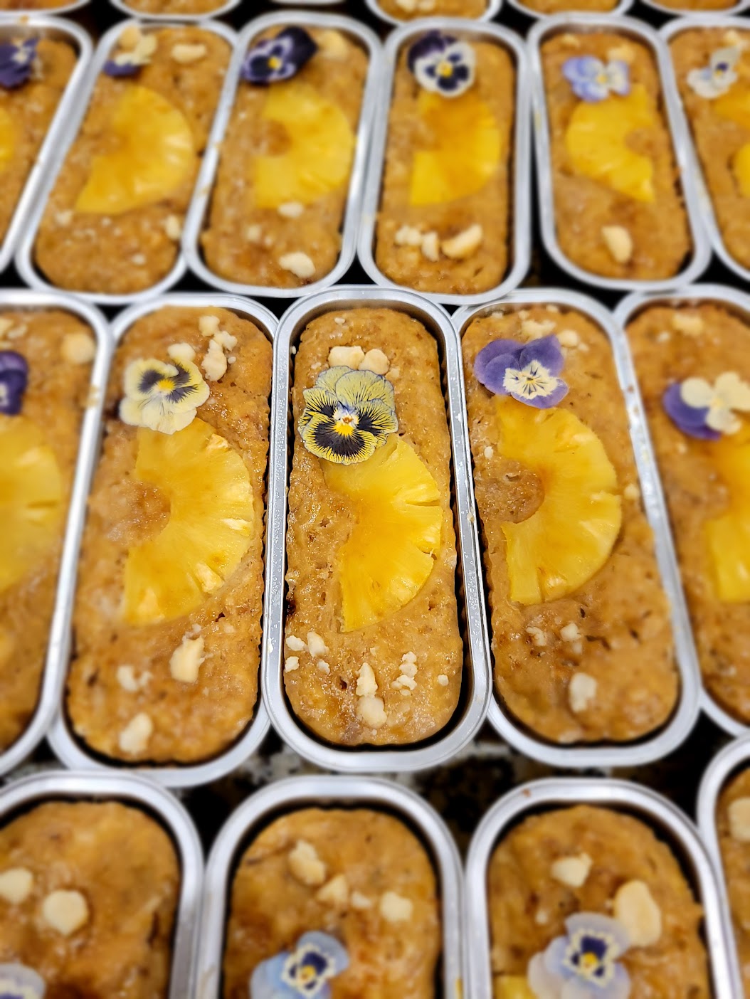 Multiple rectangular trays filled with a brown crumbly dessert topped with pineapple slices and edible flower decorations.