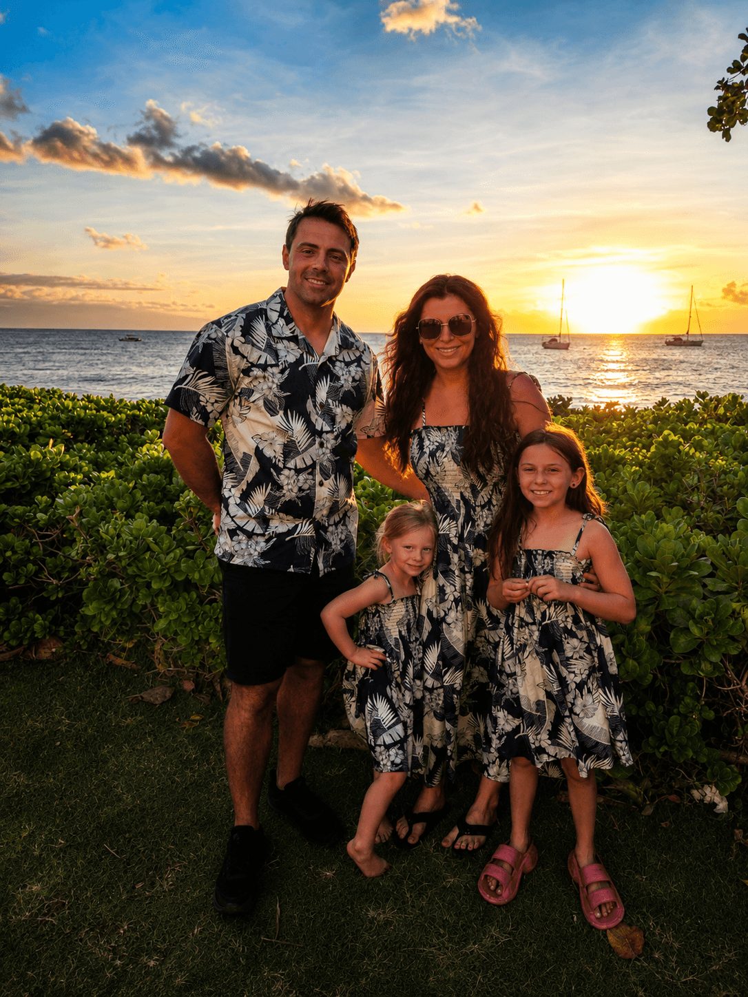 Family of four standing outdoors near the ocean at sunset, smiling, with greenery and sailboat in background.