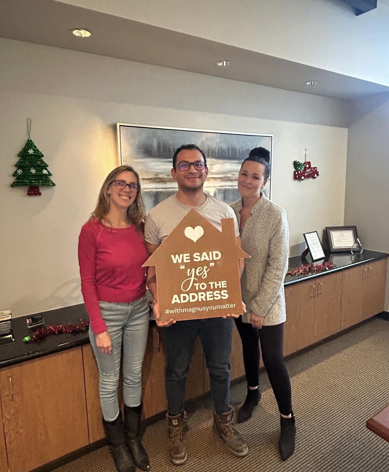 Three people standing indoors, holding a sign that says