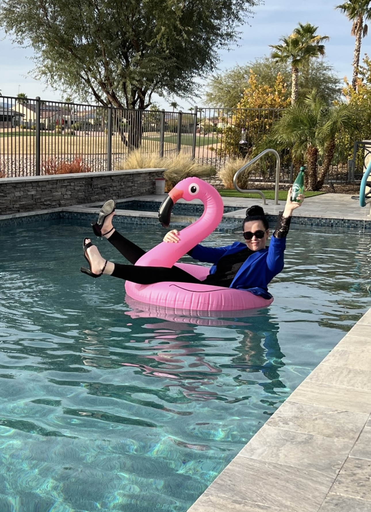 Person in sunglasses and blue outfit relaxing on a pink flamingo pool float in a swimming pool, with trees and a fence in background.